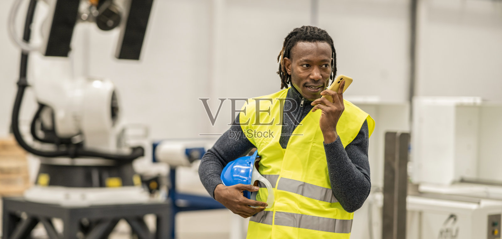 帅气的非洲男性工程师，戴着头盔，用手机看向别处。站在大型工厂坦克旁边用智能手机打电话。工作的概念照片摄影图片
