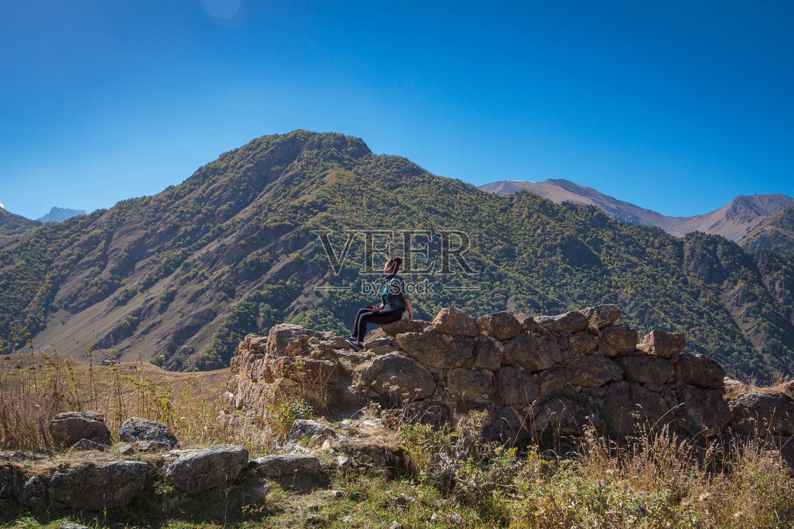 这个女孩坐在高山上的一块石头上，欣赏着大自然的景色。照片摄影图片
