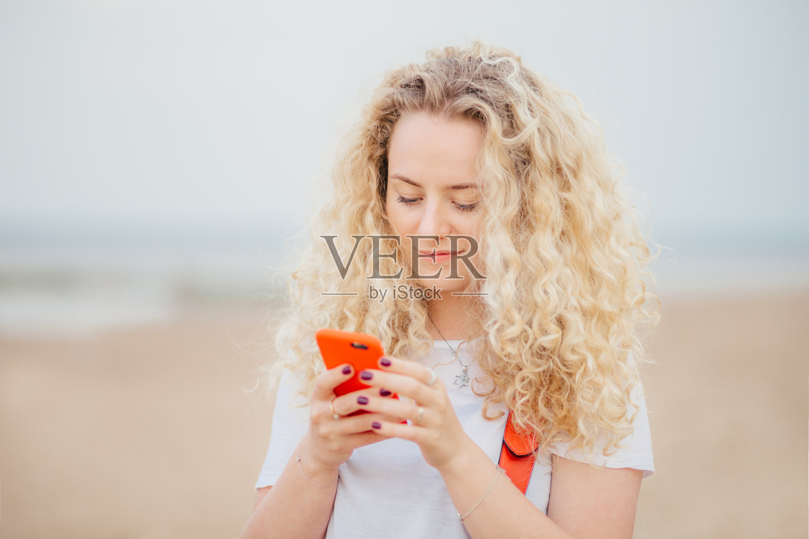 户外肖像令人愉快的卷发女性使用现代手机进行在线交流，随时保持联系，在沙滩上度假，有暑假。其他的概念照片摄影图片