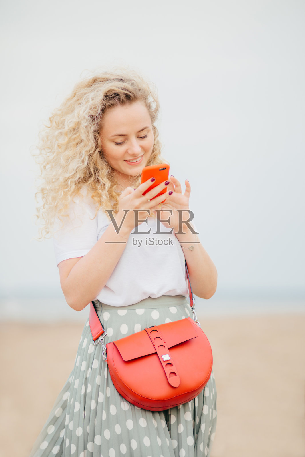 快乐漂亮的年轻女性，浅卷发，用手机浏览社交网络，穿着休闲的t恤和裙子，从男朋友那里收到愉快的信息，独自在海滩漫步照片摄影图片