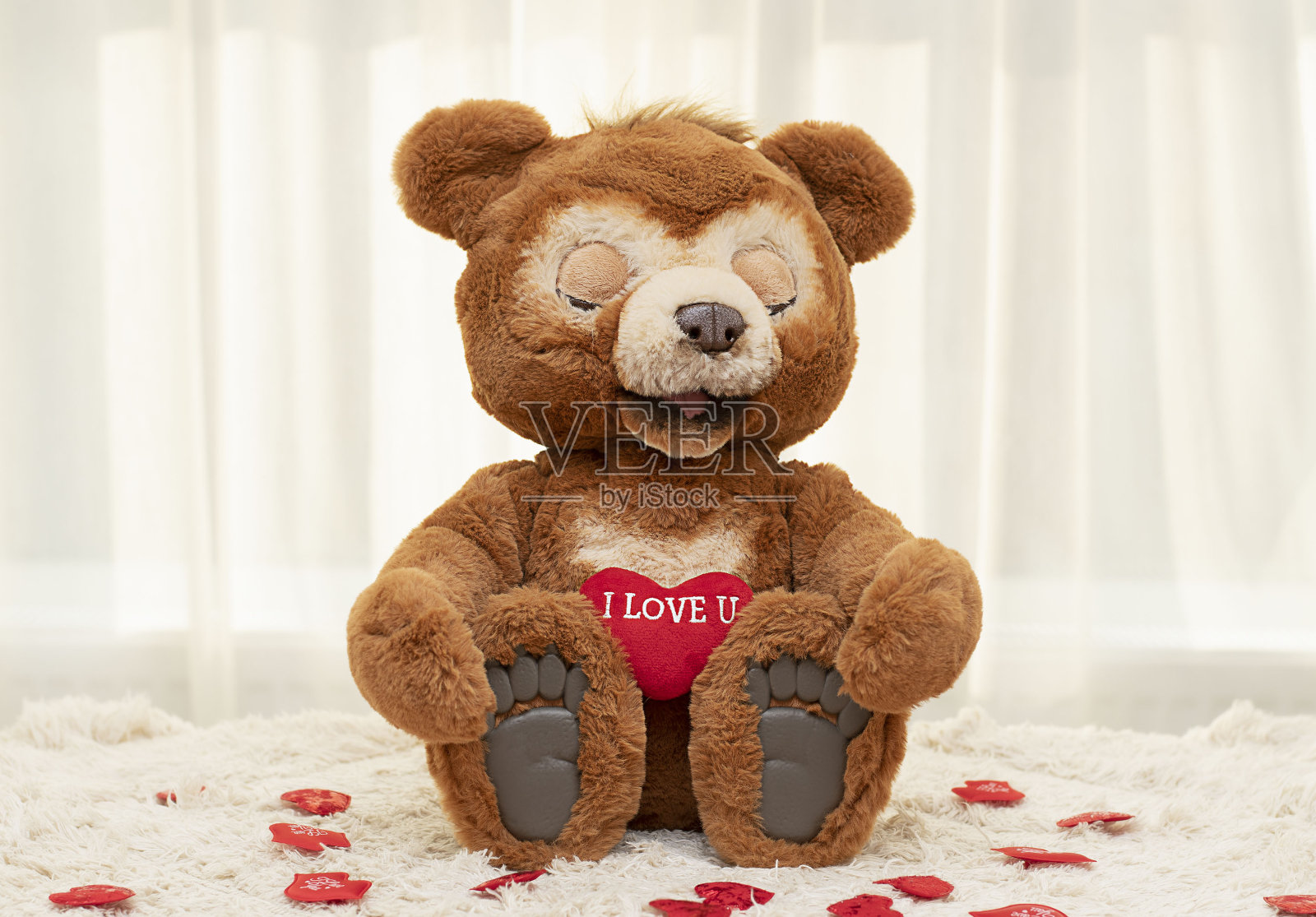 情人节的概念。一只可爱的棕色大眼睛泰迪熊坐在靠窗的地毯上，上面有一颗鲜红色的心。照片摄影图片