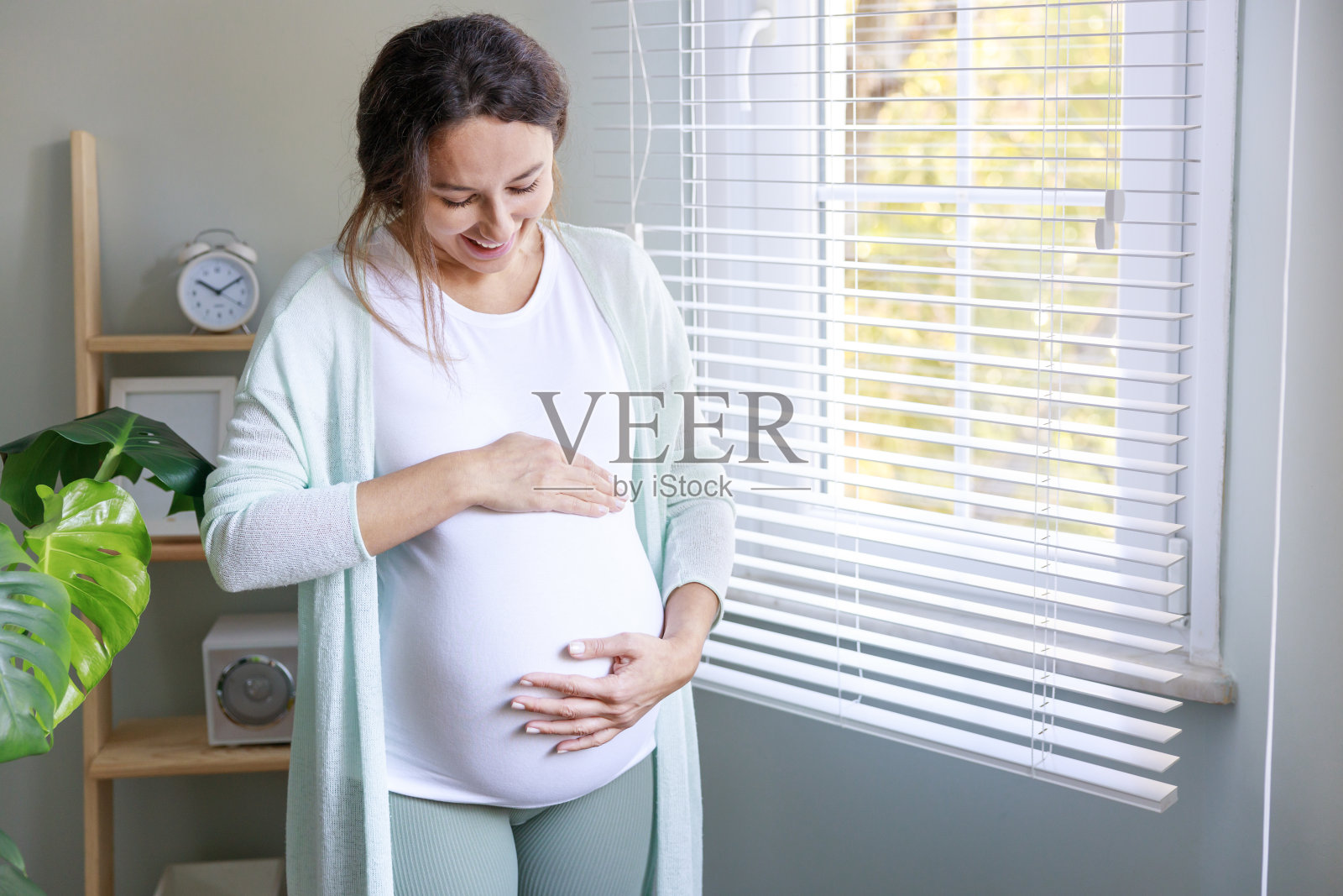 快乐的年轻孕妇摸着她怀孕的肚子，站在窗前照片摄影图片