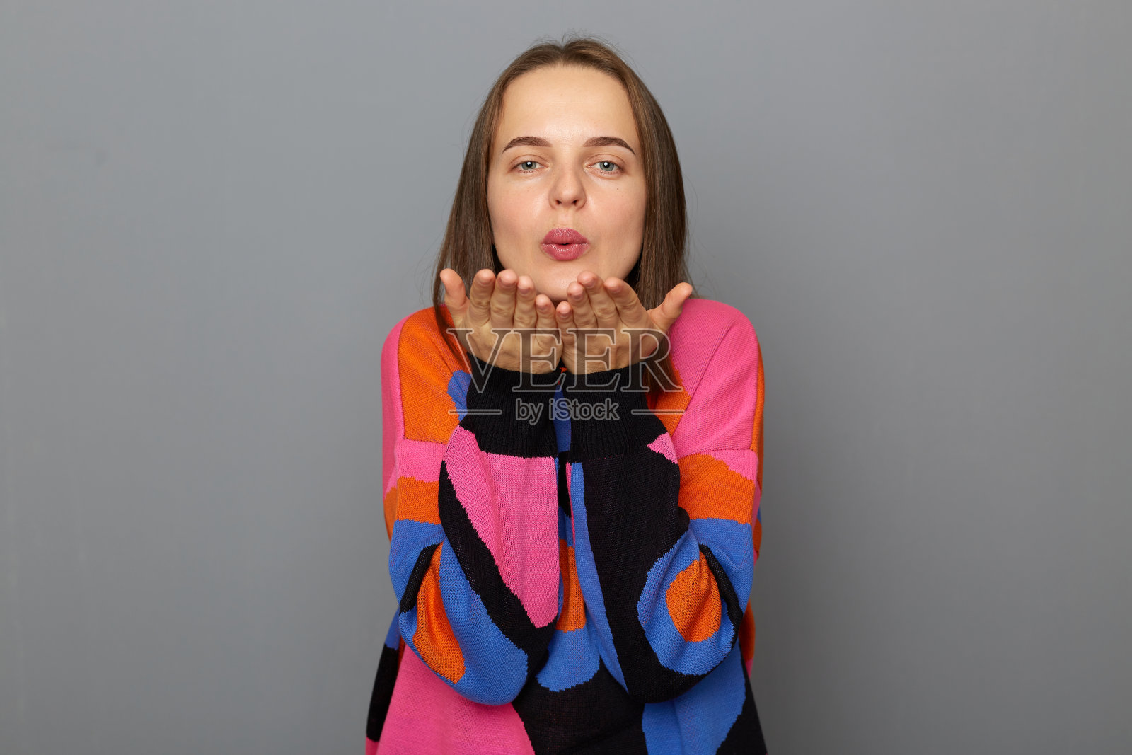 照片中愉快的迷人的年轻女子棕色头发使飞吻，保持嘴唇折叠，向亲密的人表达感情，穿着套头衫站在灰色背景上孤立。照片摄影图片