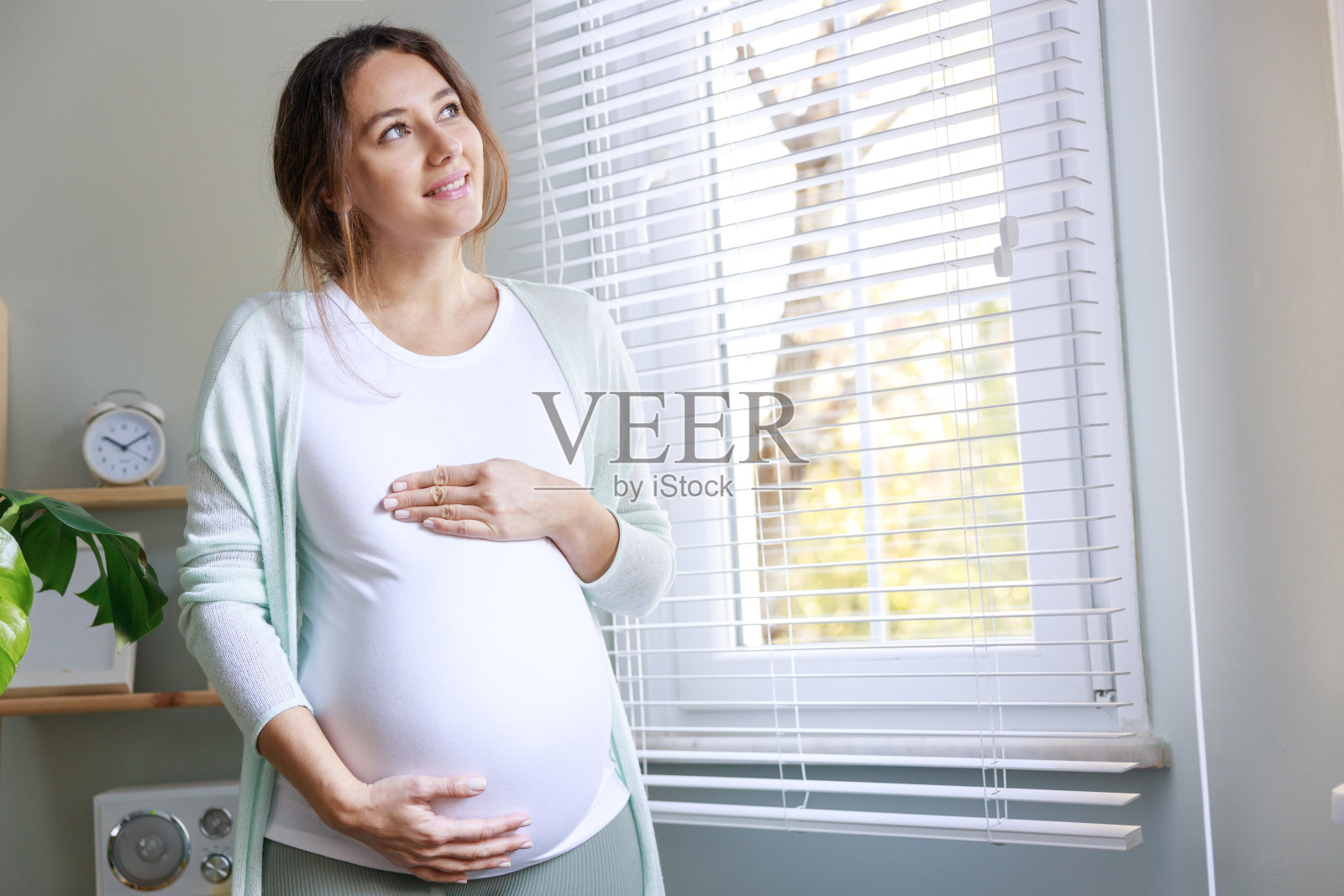 快乐的年轻孕妇摸着她怀孕的肚子，站在窗前照片摄影图片