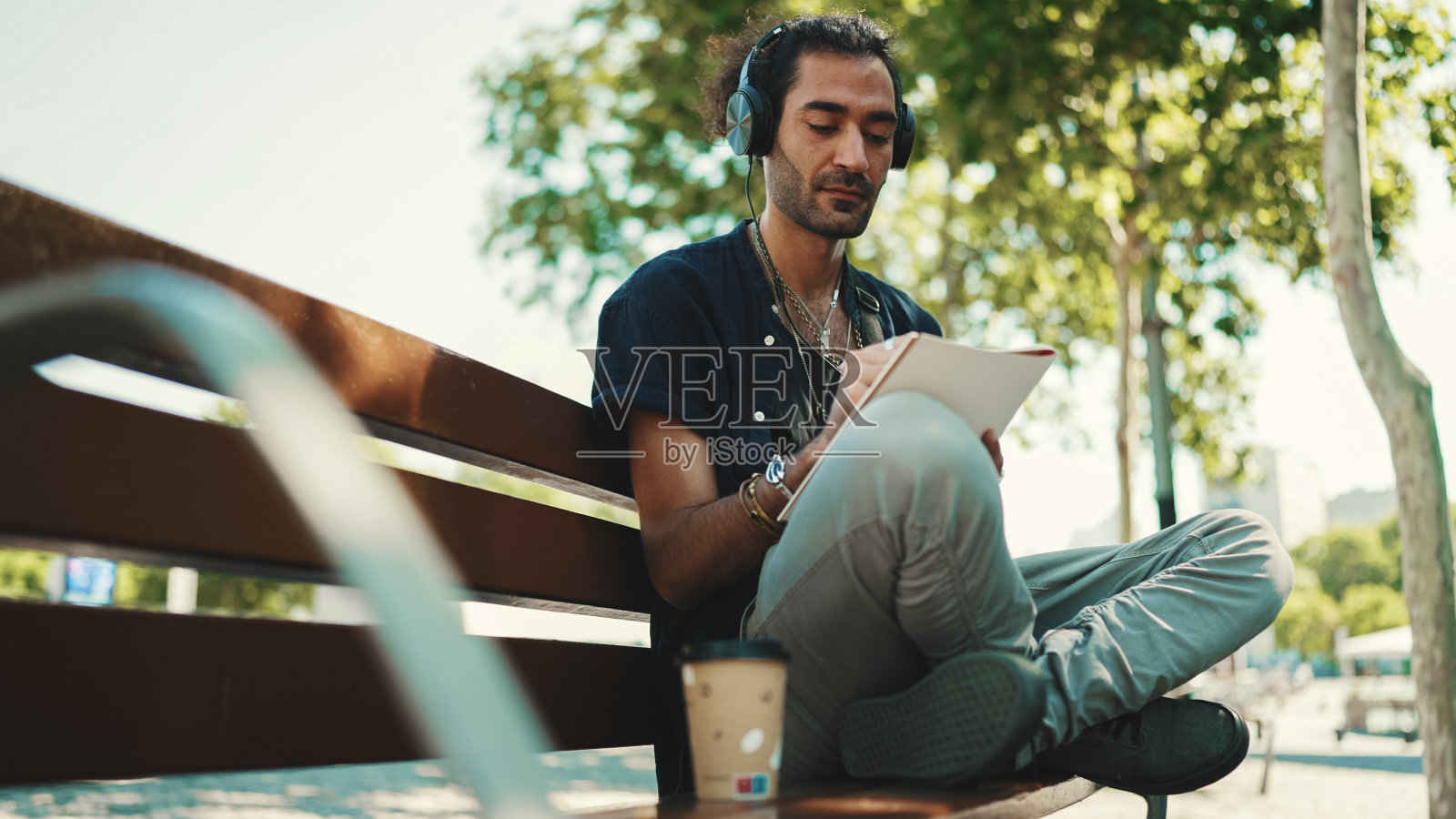 年轻的意大利小伙子梳着马尾和胡茬，戴着耳机坐在街边长凳上听音乐，在城市风景的背景下用笔在纸上画草图照片摄影图片