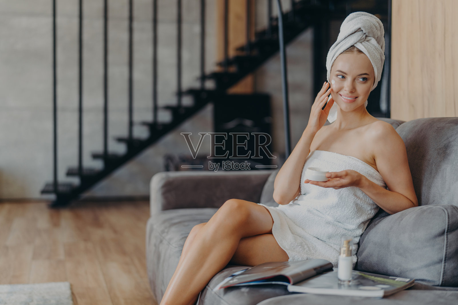 漂亮微笑的欧洲女人在脸上涂面霜，拿着一罐化妆品，照顾皮肤和肤色，在舒适的沙发上摆姿势，杂志对着楼梯的背景。照片摄影图片