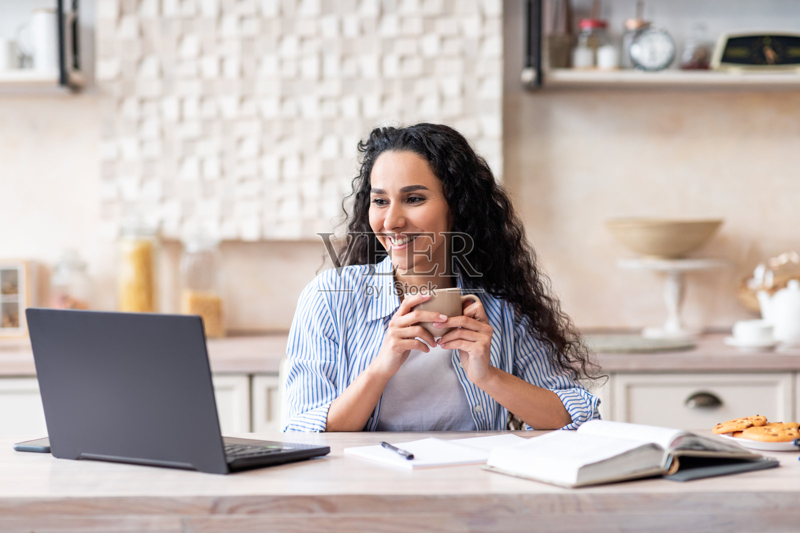 现代职员居家办公。快乐的女人拿着杯子，在厨房的桌子上用笔记本电脑工作照片摄影图片