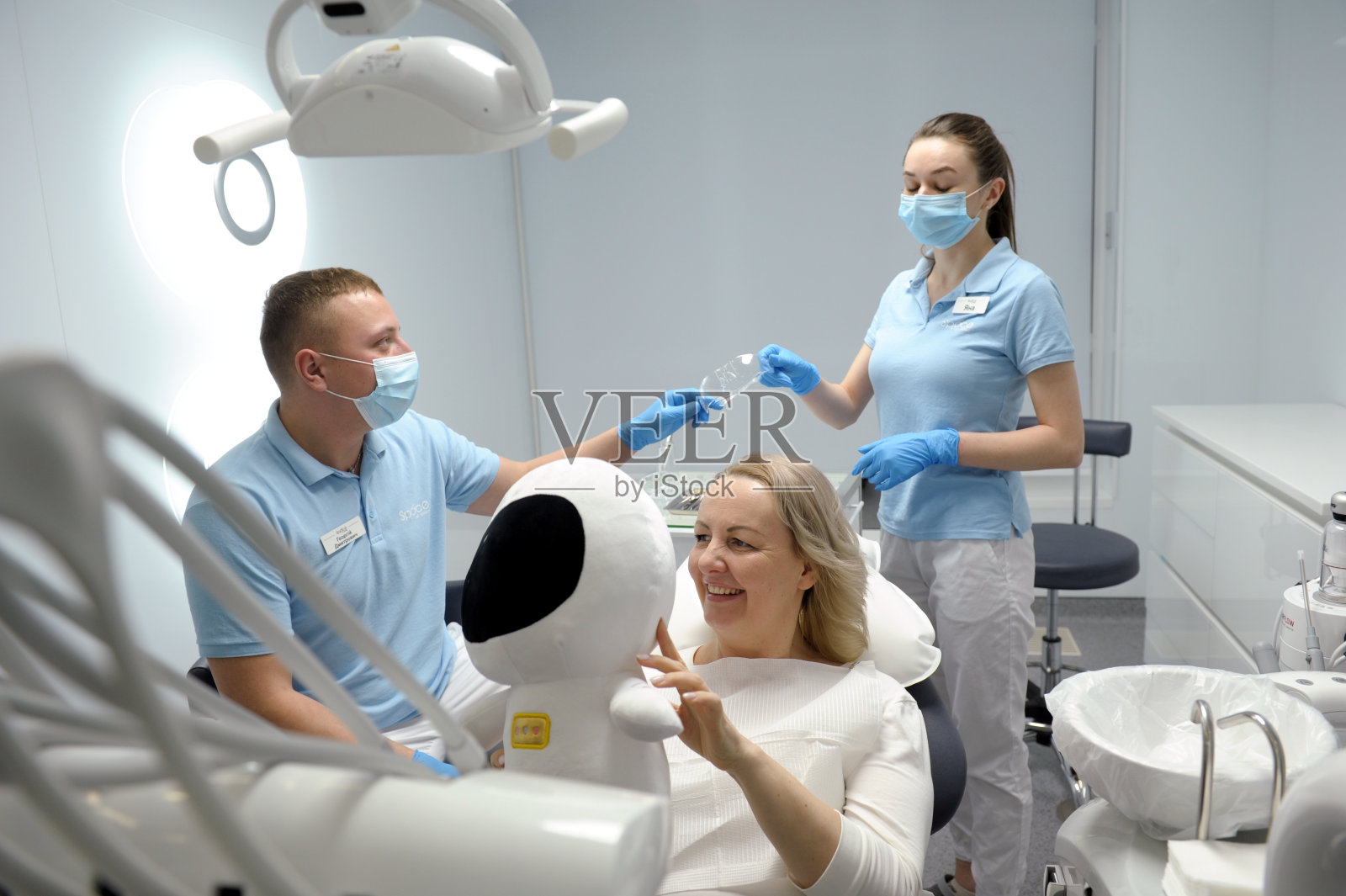 牙科诊所中年妇女坐在椅子上，宇航员柔软的玩具微笑着漂亮地向孩子展示，坐着是安全的，害怕牙科儿童护士递给医生护目镜照片摄影图片