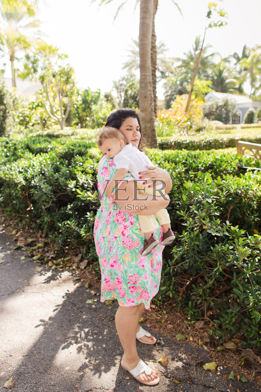 一位快乐的31岁古巴母亲，棕色卷发，穿着五彩缤纷的春天连衣裙，抱着她温暖的男婴，穿着白色短袖衬衫，浅黄色裤子和棕色鞋子，在佛罗里达州棕榈滩的湖泊小径上平静地散步照片摄影图片