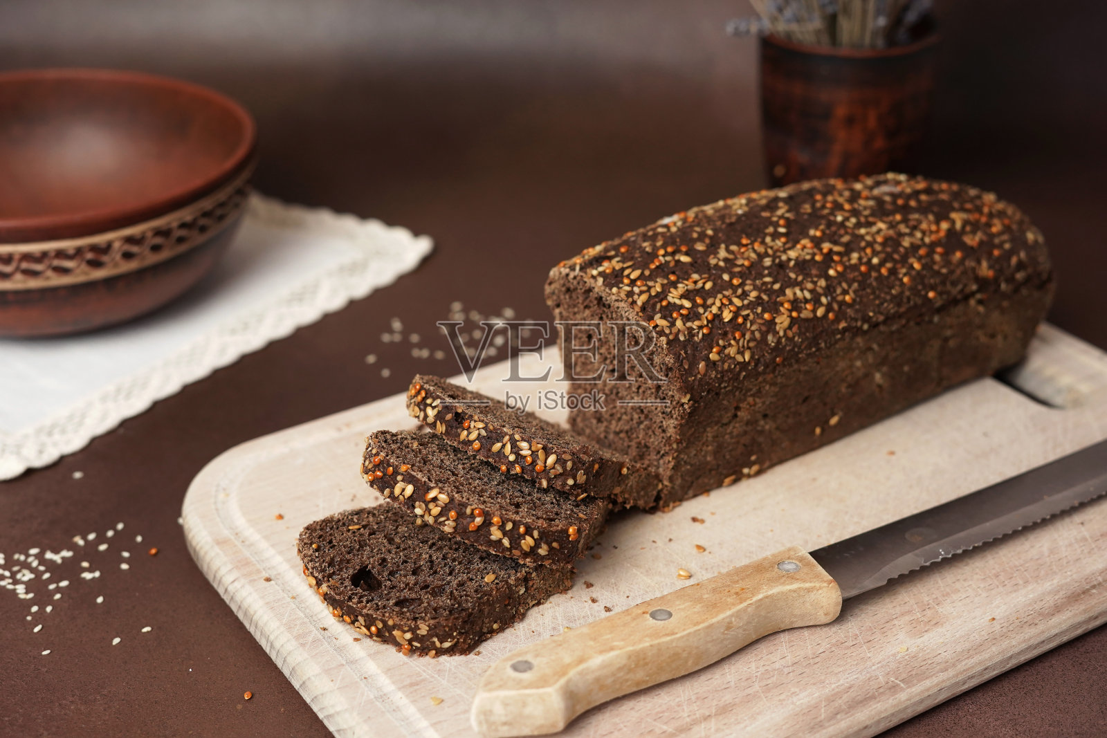 黑面包配麦片，切在棕色背景的木砧板上。面包旁边放着一把刀，陶制的盘子，叉子，勺子和白色的棉布桌布照片摄影图片