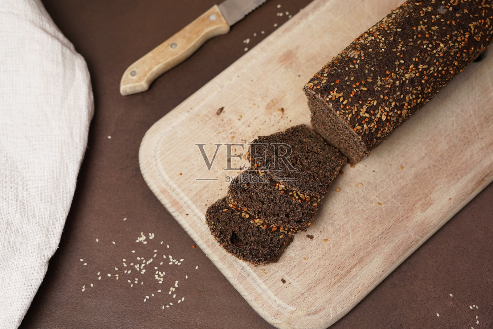 黑面包和谷物的俯视图，在棕色背景的木砧板上切成薄片。面包旁边放着一把刀和一块白色的棉布桌布照片摄影图片