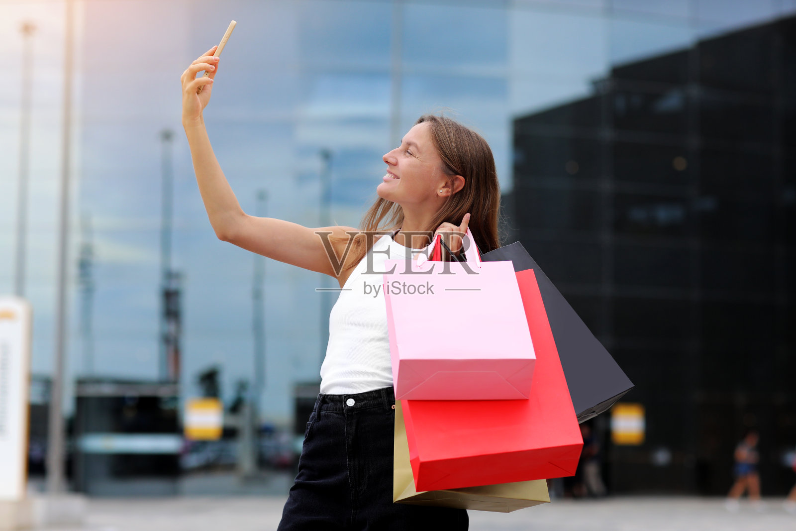 挎着购物袋和智能手机的年轻博主微笑着在购物中心附近的社交网络上与粉丝交流。消费主义概念，销售，丰富生活，虚拟生活。黑色星期五照片摄影图片