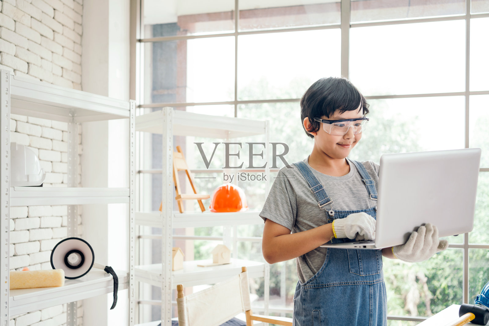 一个快乐的亚洲木匠男孩戴着护目镜和工作手套，在家庭工作室的笔记本电脑上工作。建筑工程及维修。木工和家庭观念。照片摄影图片