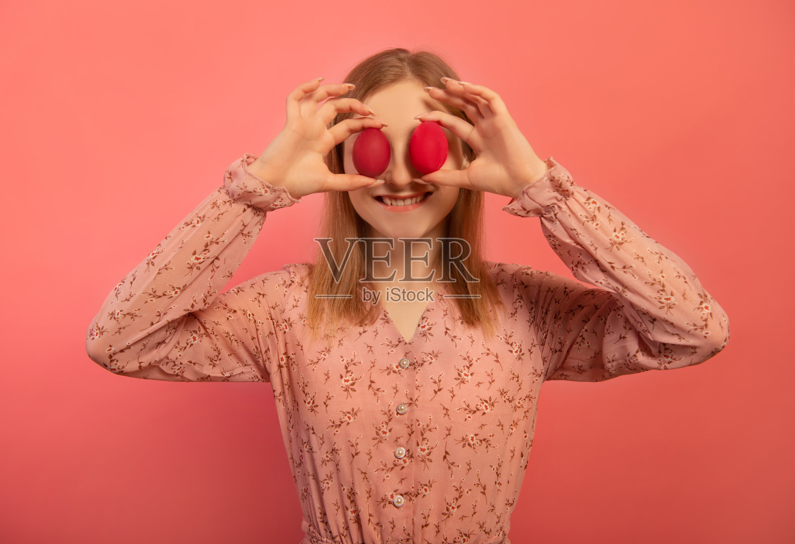 快乐的微笑的年轻女子拿着两个红色的复活节彩蛋或两个红色的复活节彩蛋在眼睛上，使快乐的表情孤立在粉红色的背景上。

快乐的女孩庆祝复活节假期。照片摄影图片