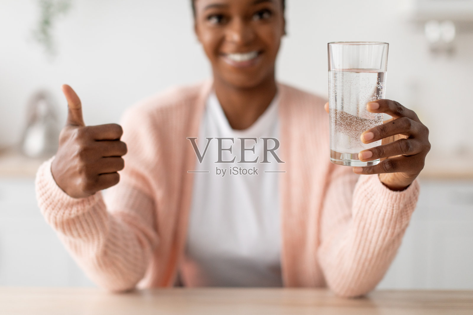 快乐的年轻非裔美国女士拿着一杯干净的水，在极简主义厨房里竖起大拇指照片摄影图片