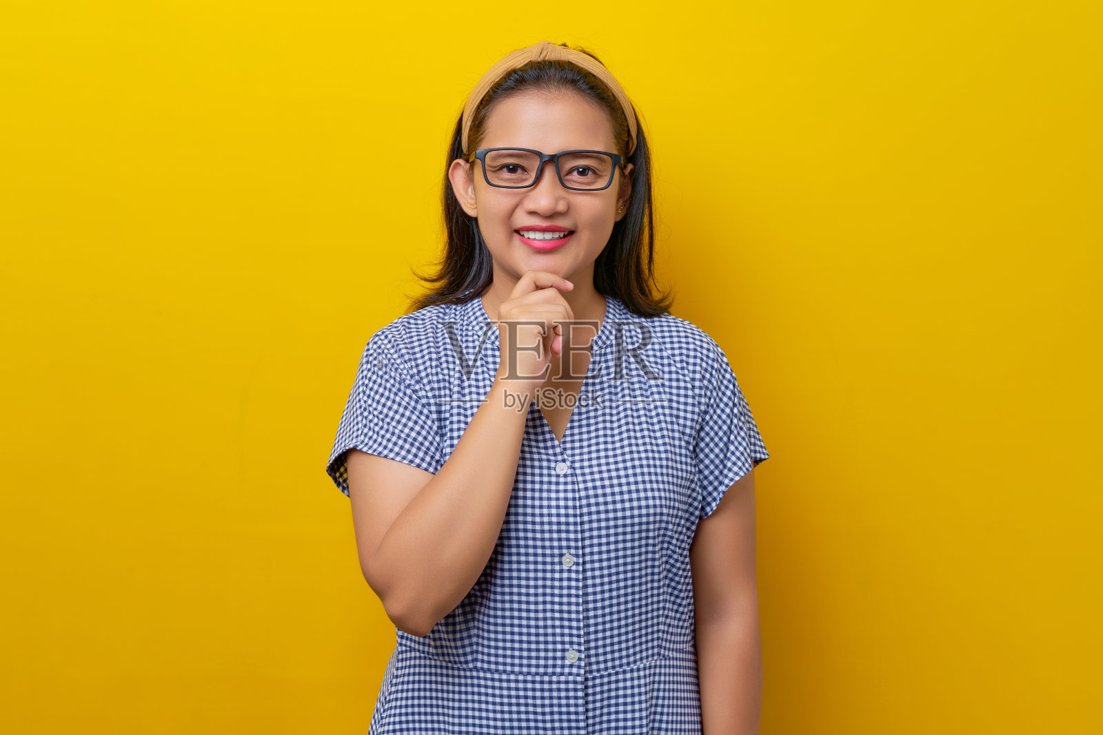 身穿带眼镜的格子连衣裙的年轻亚洲女子微笑着看着镜头，抚摸着下巴，想着被黄色背景隔离的offer。人的生活方式概念照片摄影图片