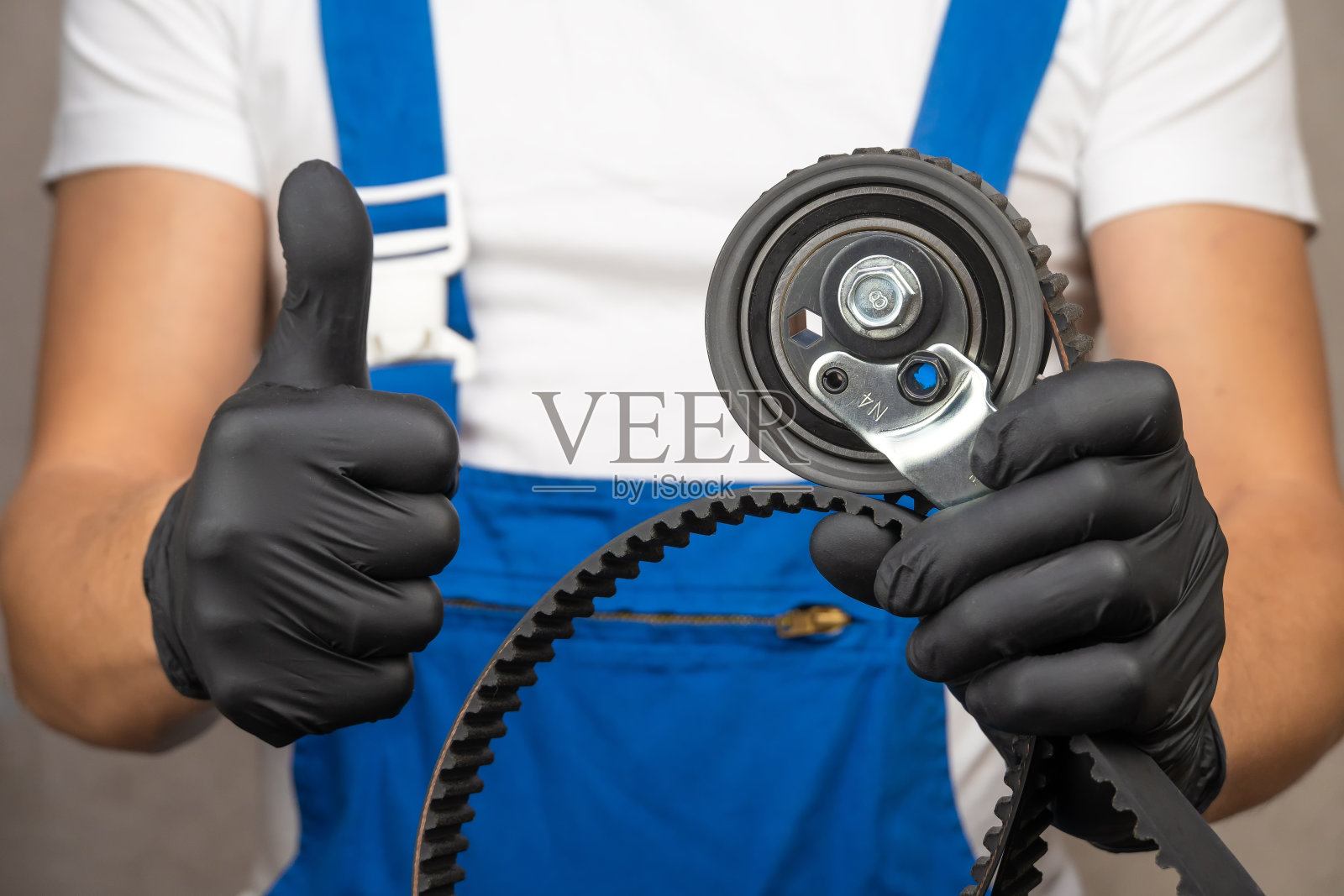 身穿蓝色连体服的汽车修理工戴着黑色手套，手里拿着发动机配气机构的滚轮皮带张紧器和皮带。汽车发动机更换皮带的概念。显示班级标志照片摄影图片