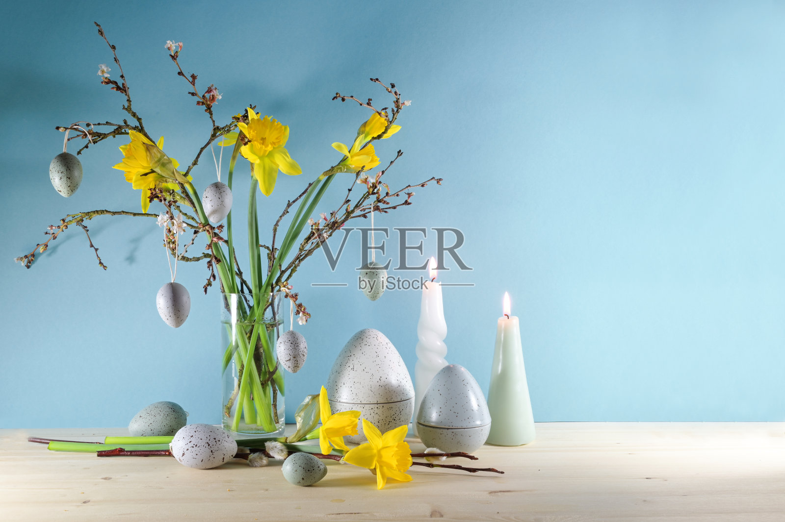 季节性的节日装饰有蜡烛，一束水仙花和春天的树枝在一个玻璃花瓶和人造灰色复活节彩蛋在一个轻木板上靠着蓝色的墙壁，复制空间照片摄影图片