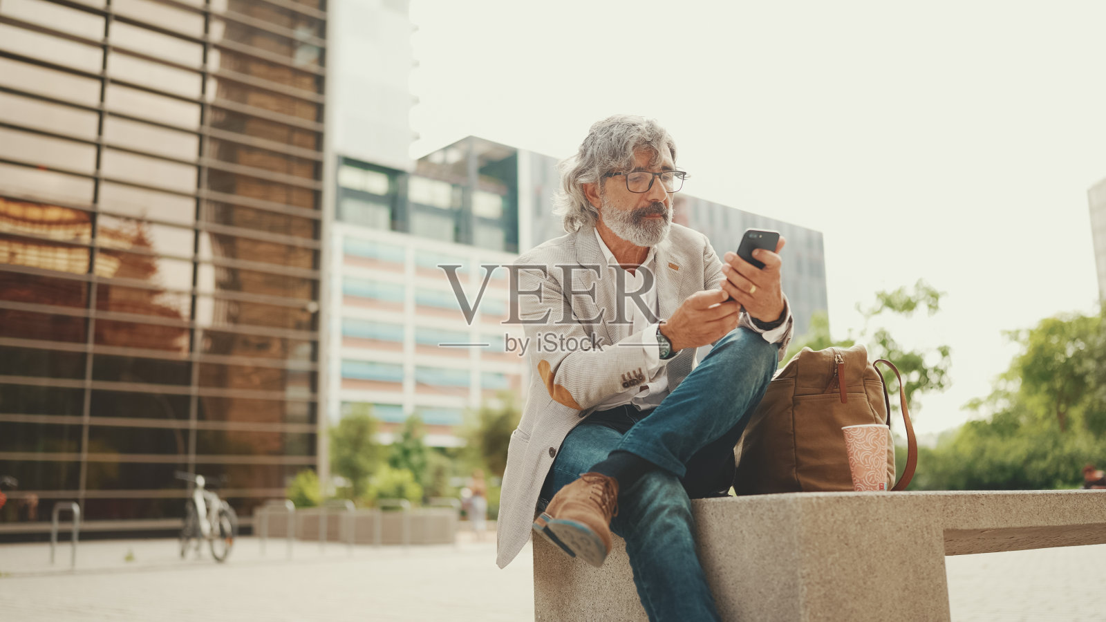 留着胡子，戴着眼镜，穿着灰色夹克的成熟商人正在使用手机。一位中年经理坐在办公室外面，用智能手机浏览信息照片摄影图片