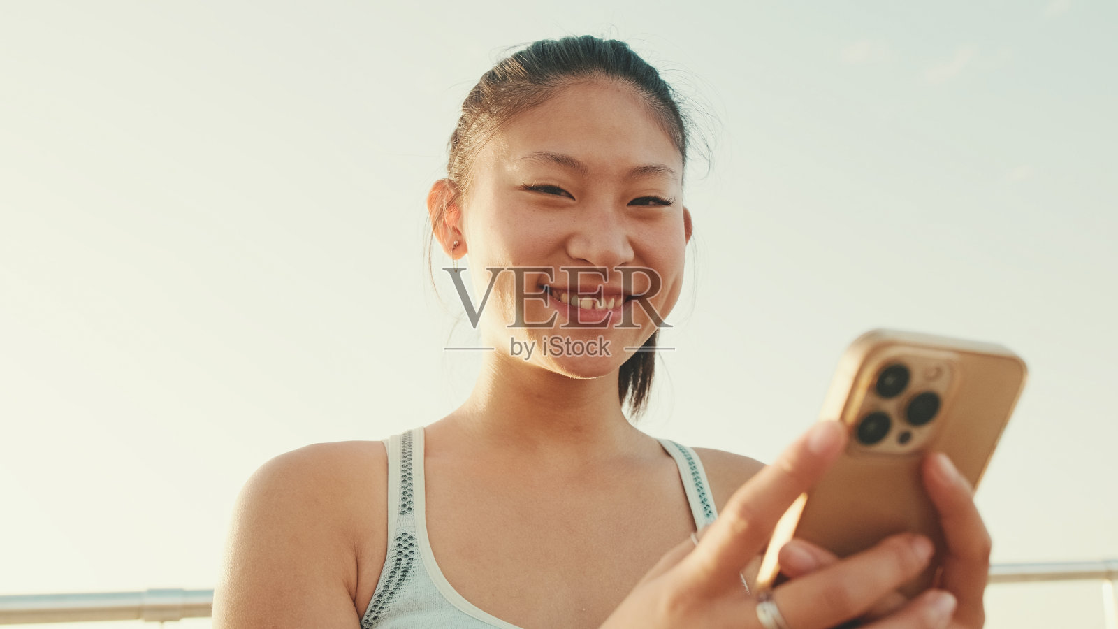在晨光中，站在现代建筑背景的海滨，微笑的亚洲女孩在运动上衣使用手机照片摄影图片