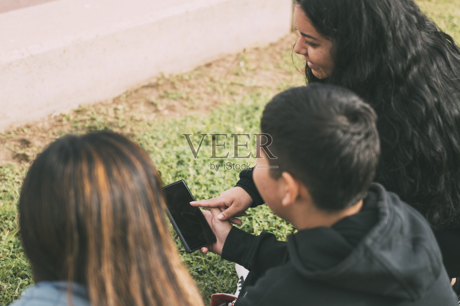 从后面拍摄的照片显示，这群人坐在草地上，看着其中一个年轻人拿着的智能手机照片摄影图片