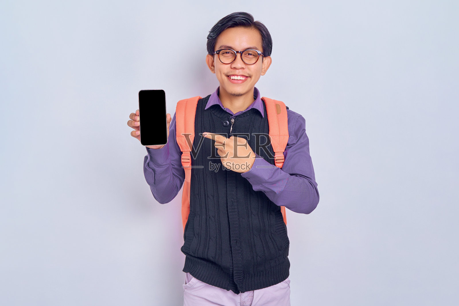 开朗的年轻亚洲学生，穿着休闲服装，背着背包，手指指着灰色背景上孤立的空白屏幕手机。高中大学学院概念照片摄影图片