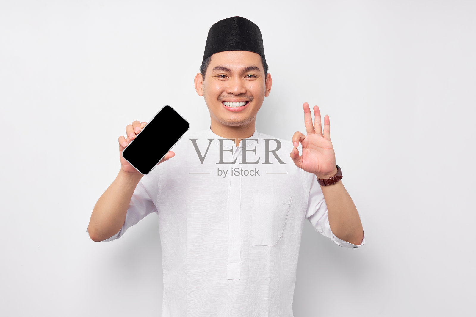 微笑的年轻亚洲穆斯林男子显示显示手机屏幕，显示ok手势孤立在白色背景。人们信仰伊斯兰教的生活方式观念。庆祝斋月并炸死了穆巴拉克照片摄影图片