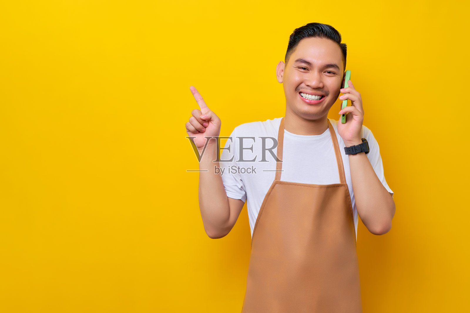 微笑的年轻亚洲男子咖啡师，穿着棕色围裙的酒吧服务员在咖啡店工作，一边说着手机，一边用手指着黄色背景上隔离的复印空间。小型企业创业照片摄影图片