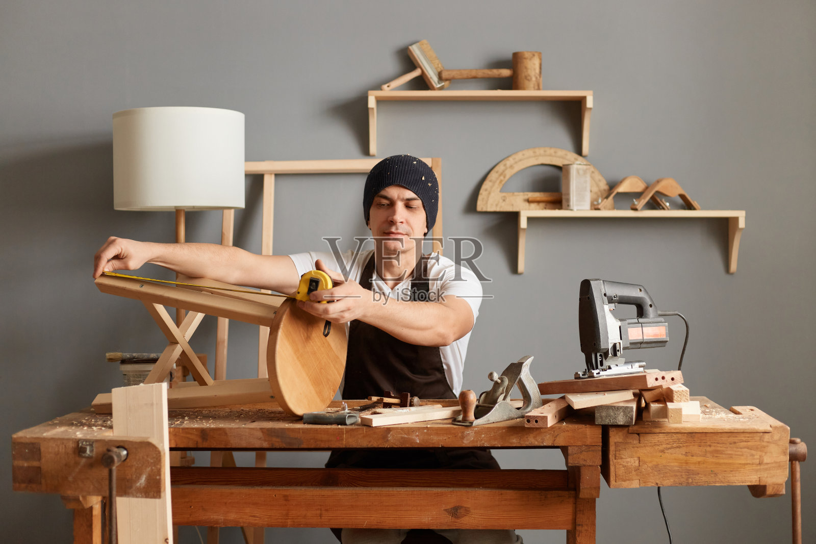严肃英俊的白人细木工肖像，戴着围裙和帽子坐在木工车间的桌子旁，测量他的车间制作的椅子腿的长度。照片摄影图片