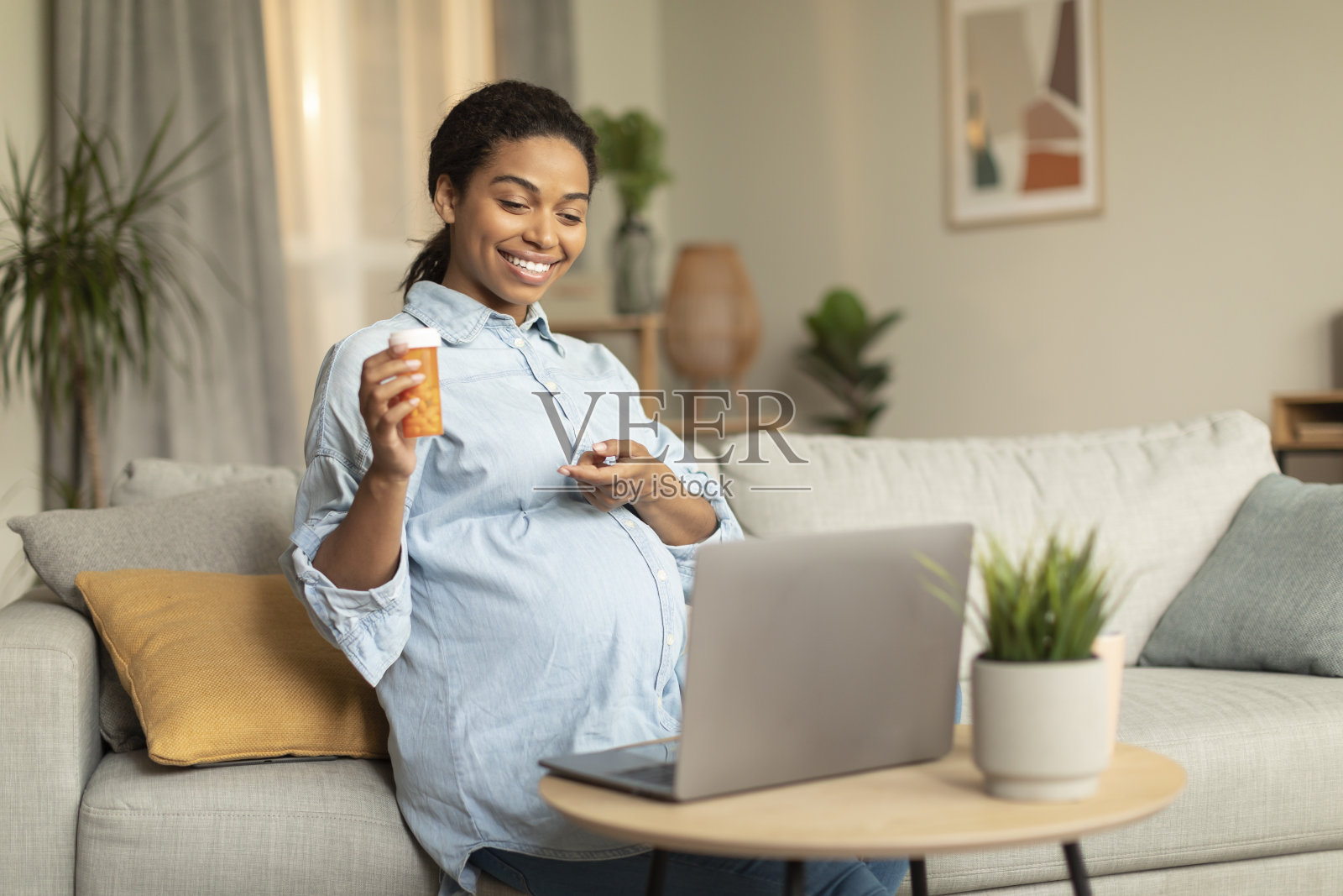 积极年轻的怀孕黑人妇女视频打电话给医生，并向笔记本电脑显示药瓶，坐在沙发上照片摄影图片