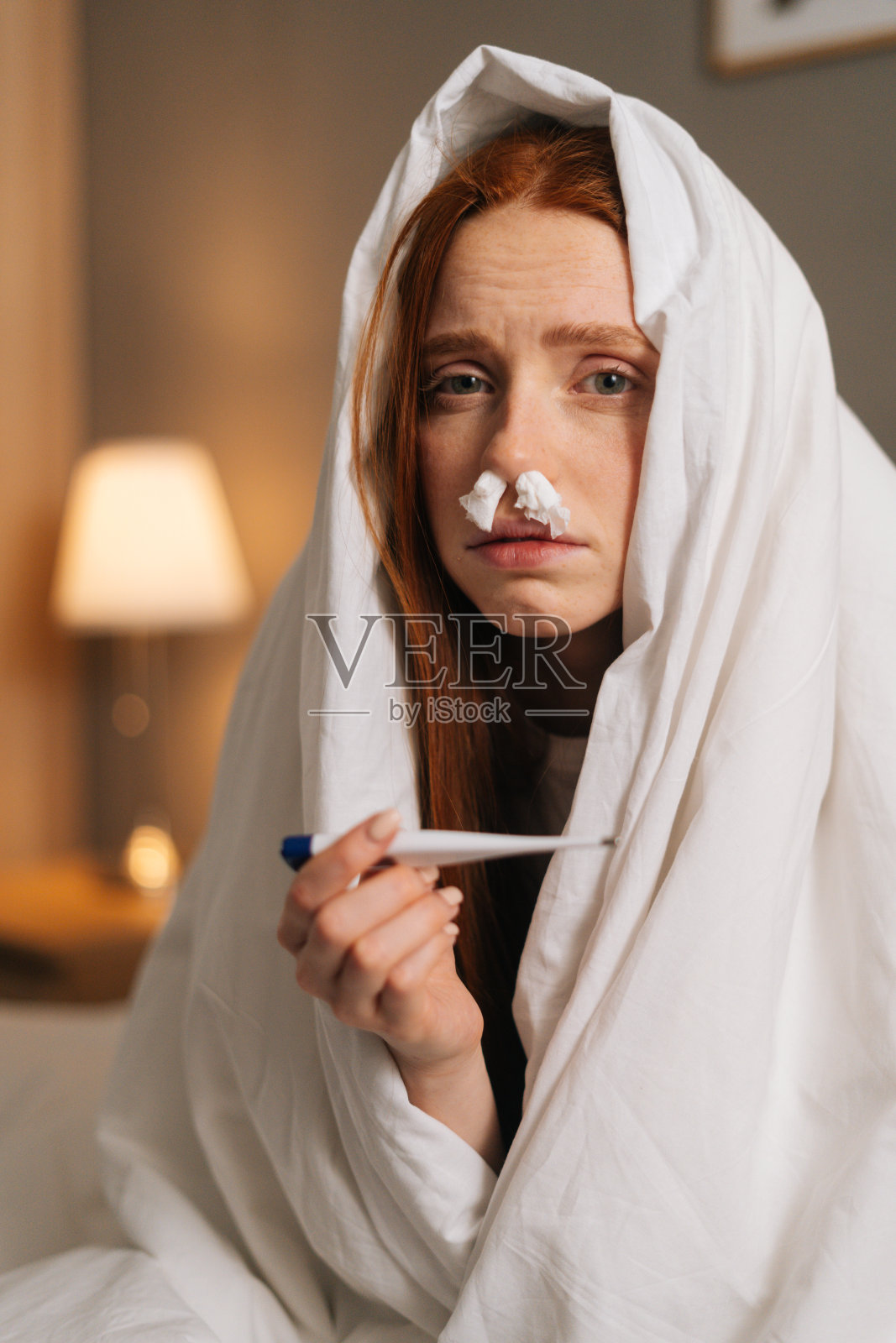 患流鼻涕病的年轻女子用纸巾插进鼻子，用体温计测量温度的垂直画像。照片摄影图片