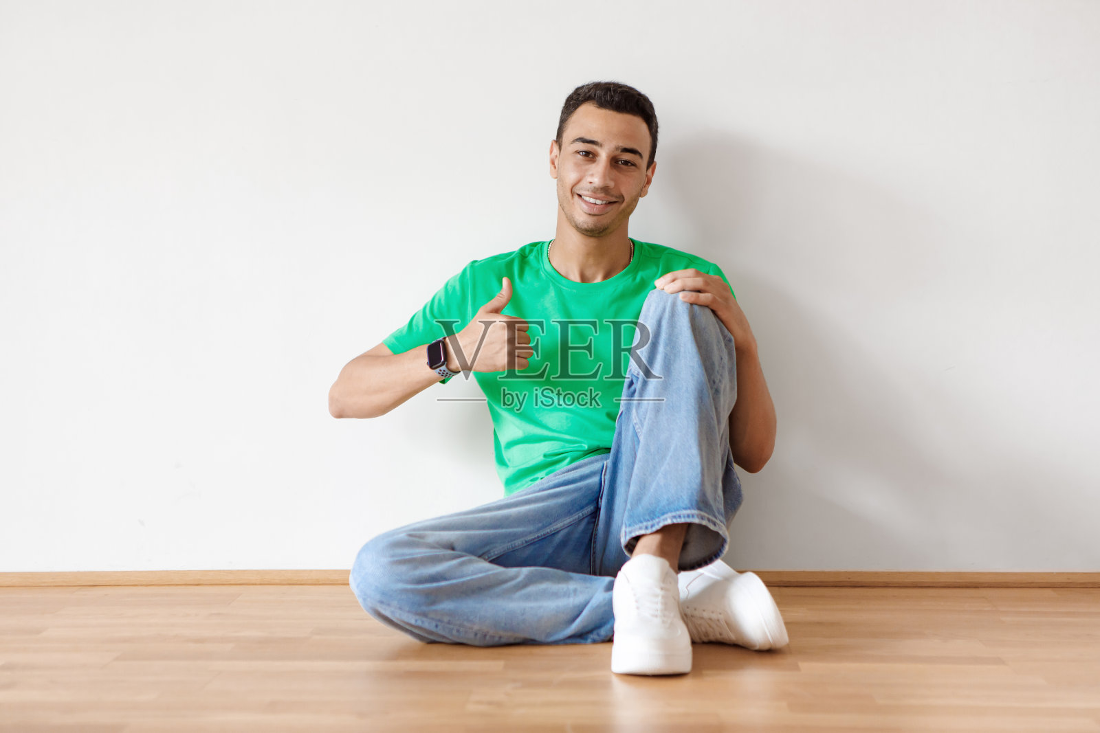 愉快的阿拉伯人坐在地板上，微笑和显示大拇指，手势的标志，对白墙的批准照片摄影图片