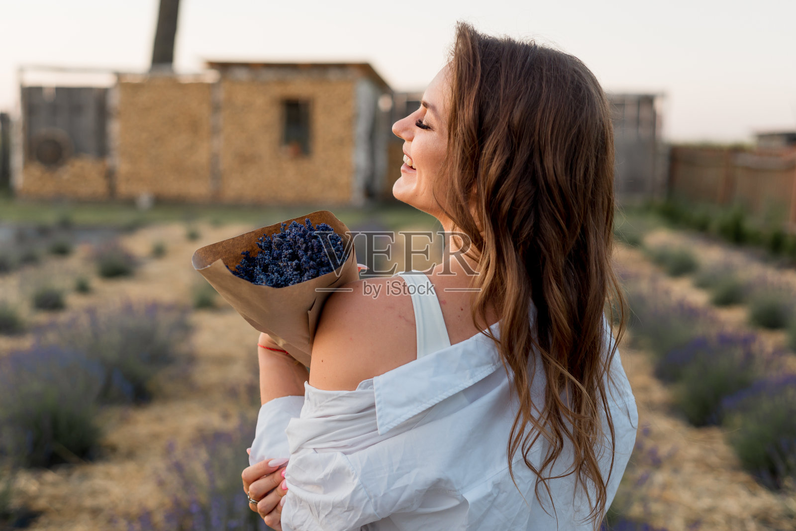 一个美丽的女孩手捧一束薰衣草站在日落时分。夏天，一个穿着白衬衫的女孩站在花丛中。休息的概念和与自然的统一。照片摄影图片