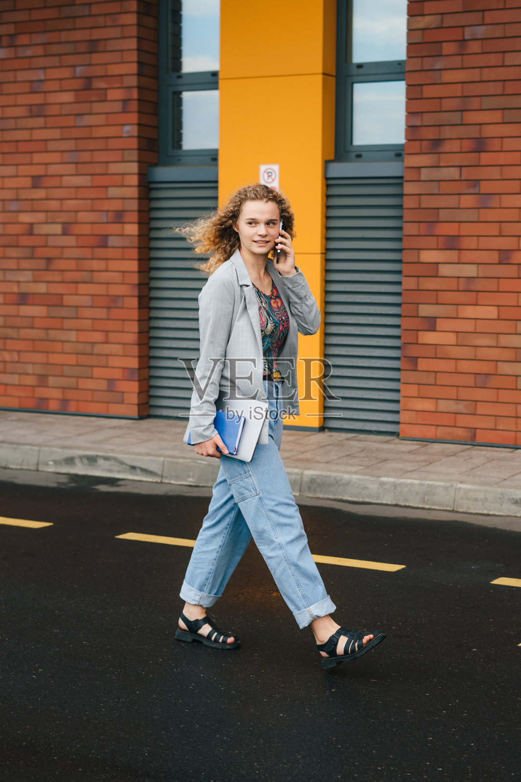 年轻的大学生穿着夹克，裤子，拿着笔记本电脑，打电话，走在办公室外的街道上。现代的生活方式。快乐的生活方式。人们生活方式的肖像。时尚风格。都市街头时尚。照片摄影图片