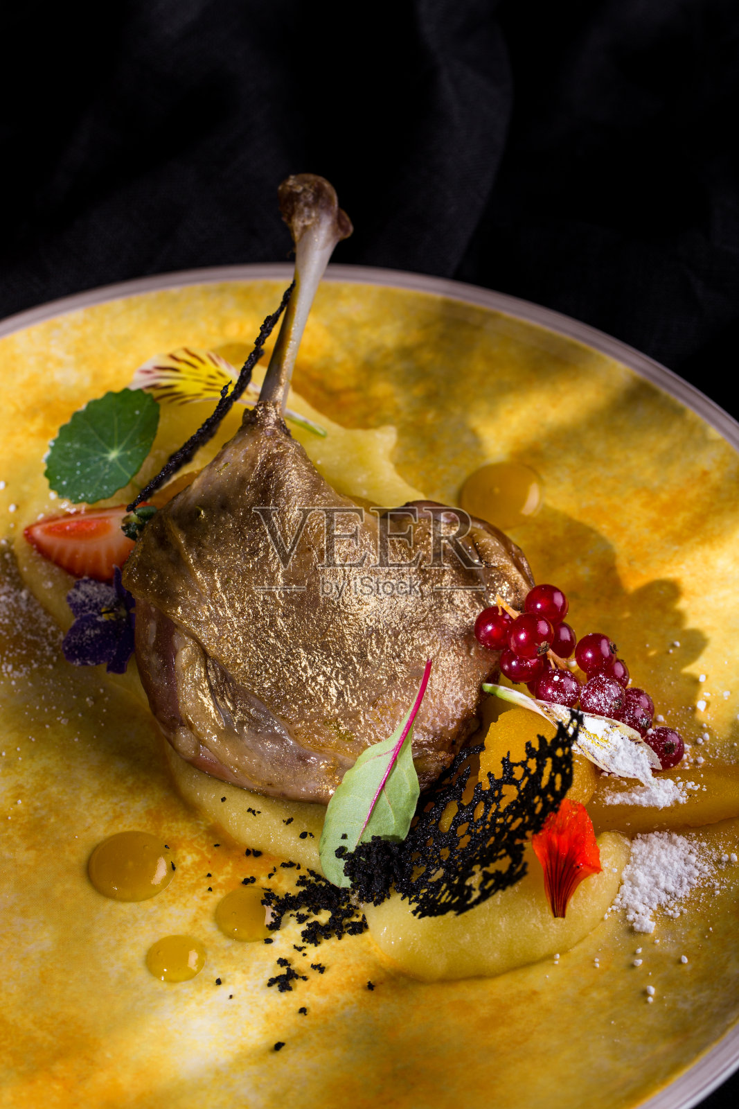 烤鸭腿带骨，金皮，红醋栗，花瓣，糖粉，草莓，土豆泥，蕾丝片，蜂蜜芥末酱放在圆形黄色盘子上，深色背景垂直方向照片摄影图片