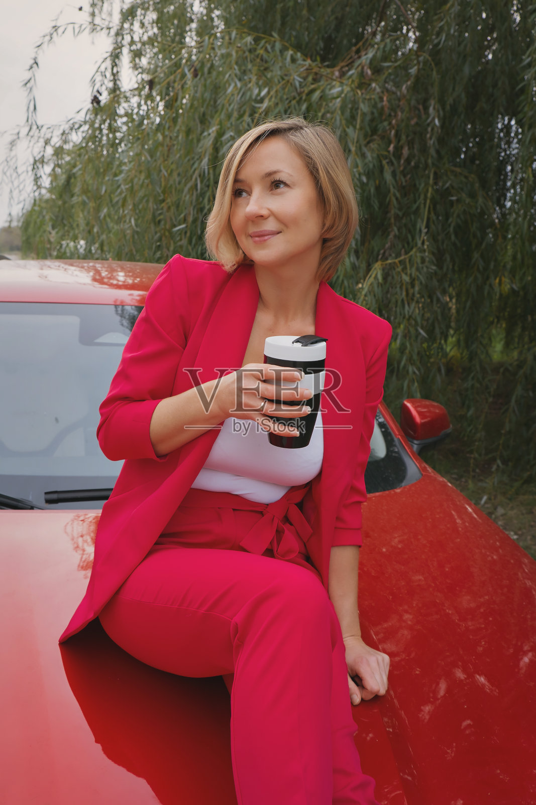 一名身着西装、坐在红色中国车里喝咖啡的女子。在线工作。自由职业，旅行，远程工作。行动自由，森林里的模特照片摄影图片