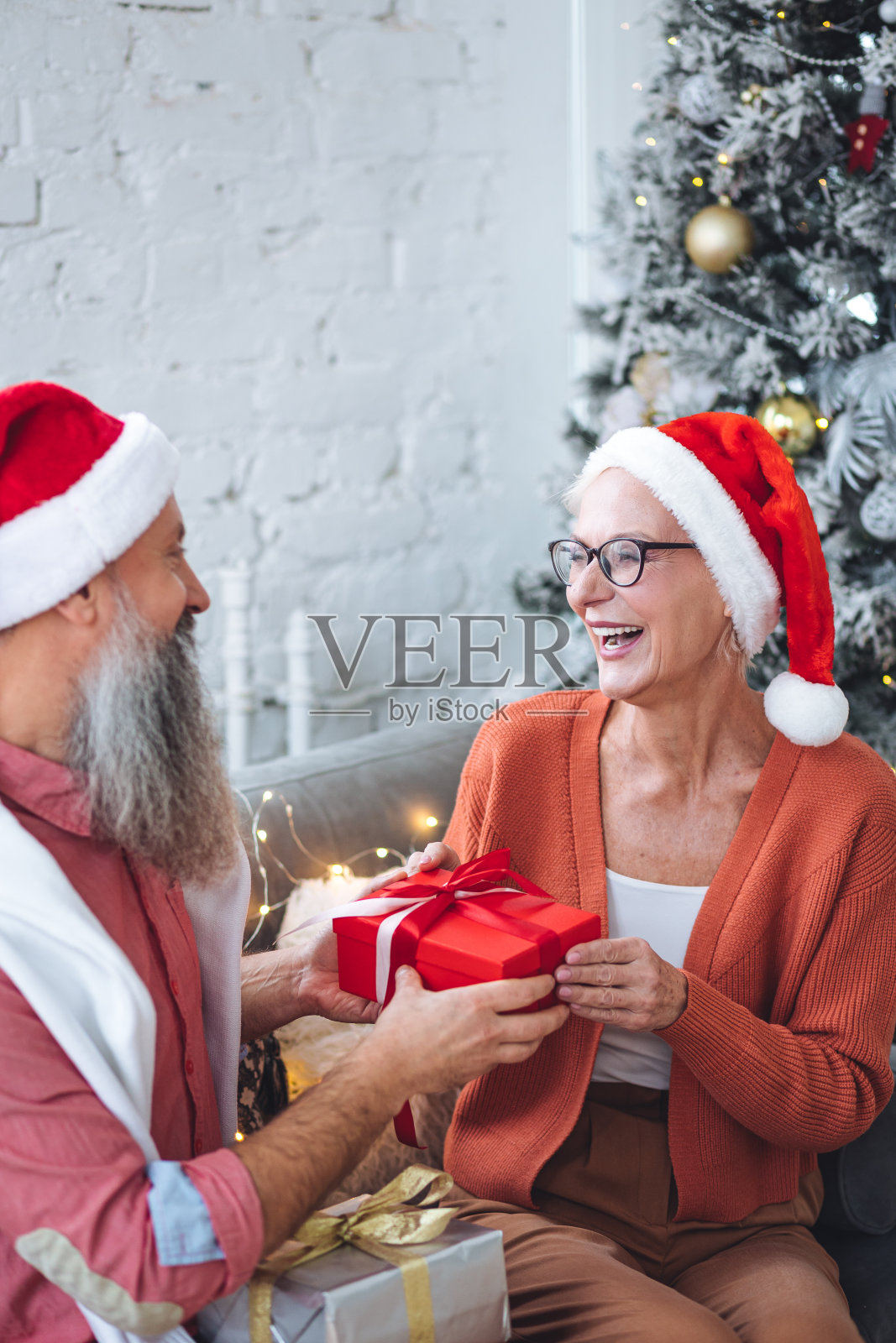 快乐的爱的老夫妇在圣诞树旁的新年晚会上玩得很开心。成熟的男人和女人微笑着，喝着香槟，交换礼物。退休人员的积极生活和休闲观念照片摄影图片