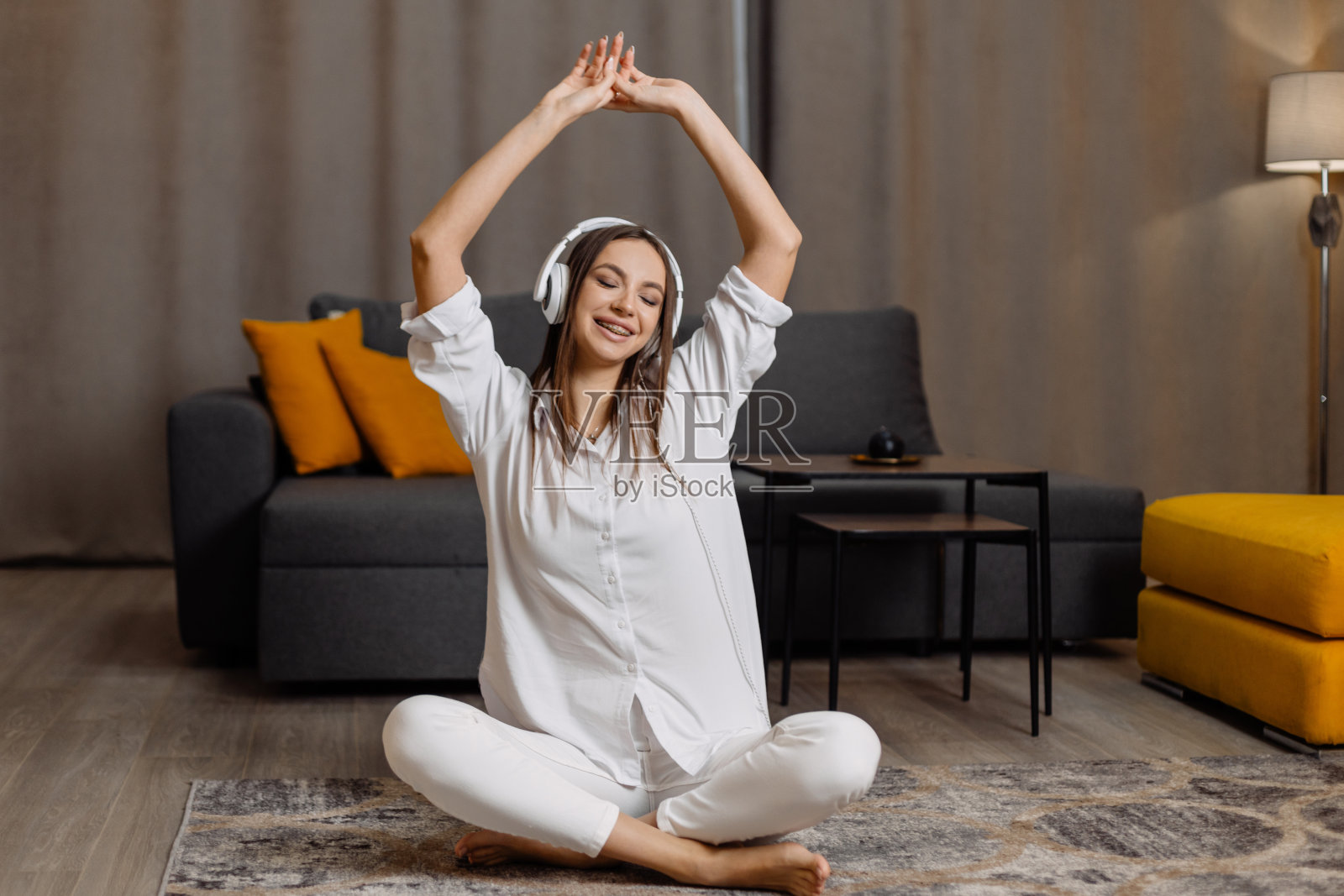 快乐的年轻白人女子坐在家里的地板上，戴着耳机用智能手机听音乐，跳舞照片摄影图片