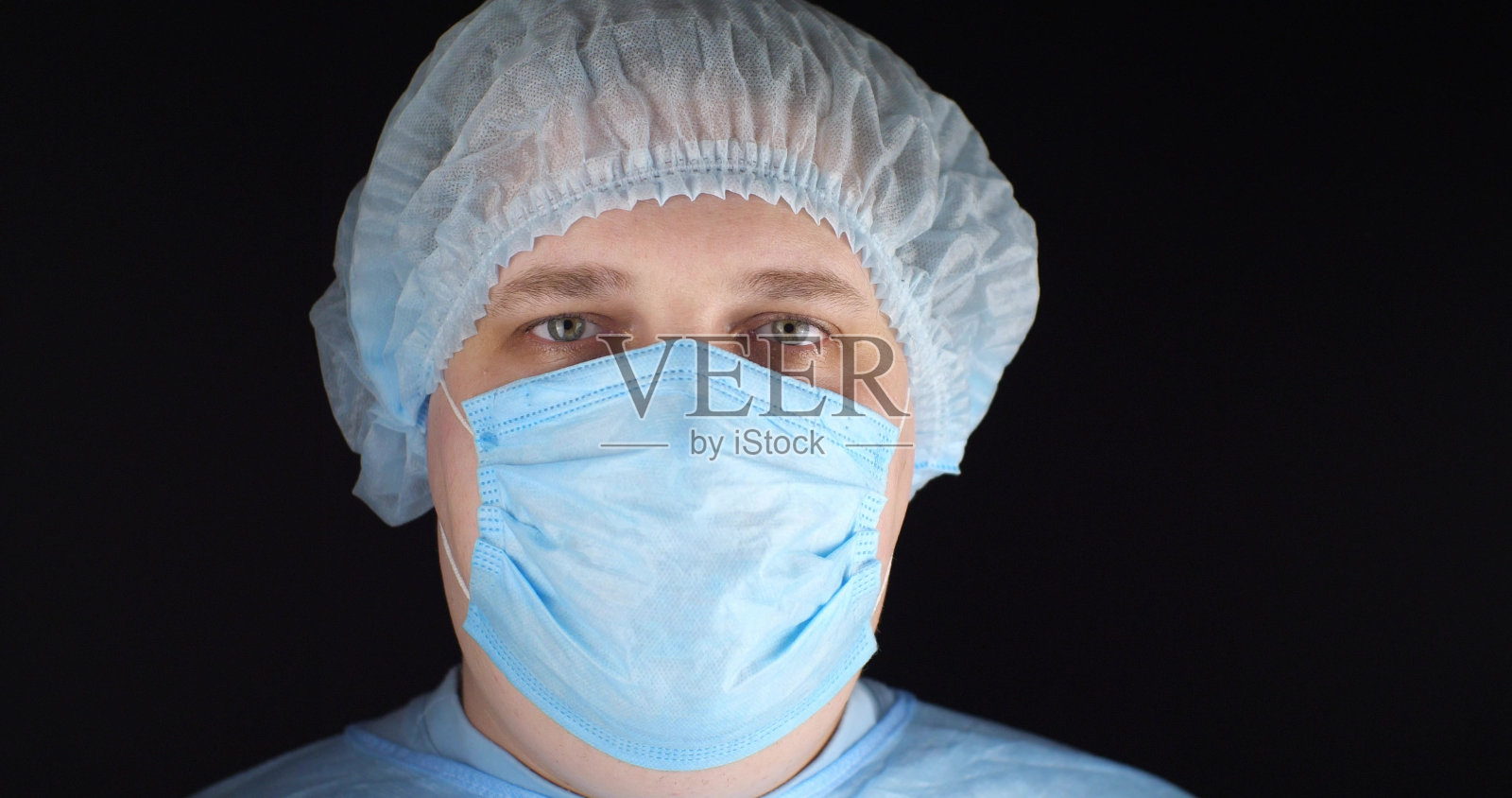 穿着蓝色制服、手术服、口罩和帽子的外科医生看着镜头。在一个孤立的黑色背景上，一名男性外科医生看着镜头。肖像,特写。照片摄影图片