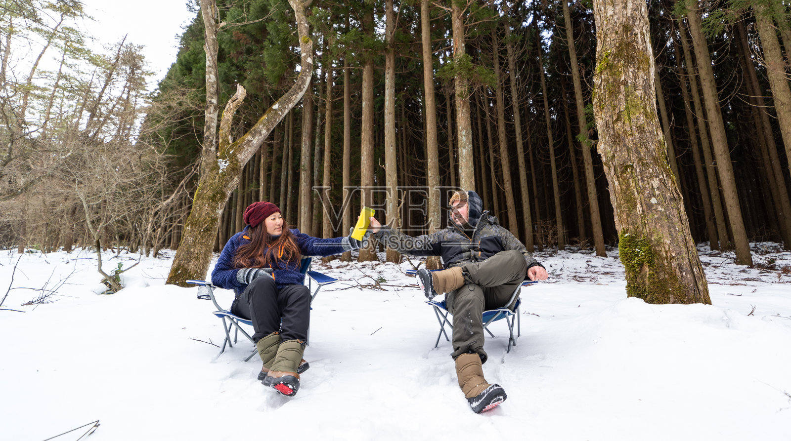一对情侣在白雪皑皑的森林风景中，在露营椅上放松，分享一瓶热饮照片摄影图片
