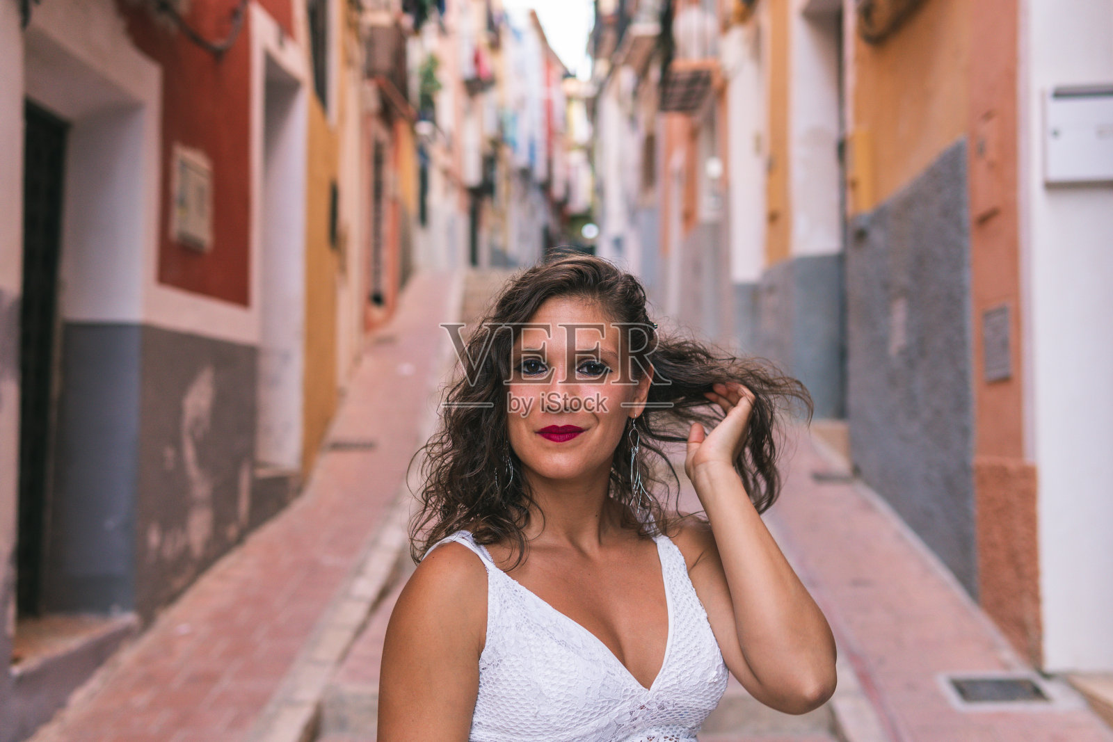 在西班牙阿利坎特五颜六色的小镇Villajoyosa，一名黑发女子在有楼梯的街道上扎头发，试图微笑。照片摄影图片