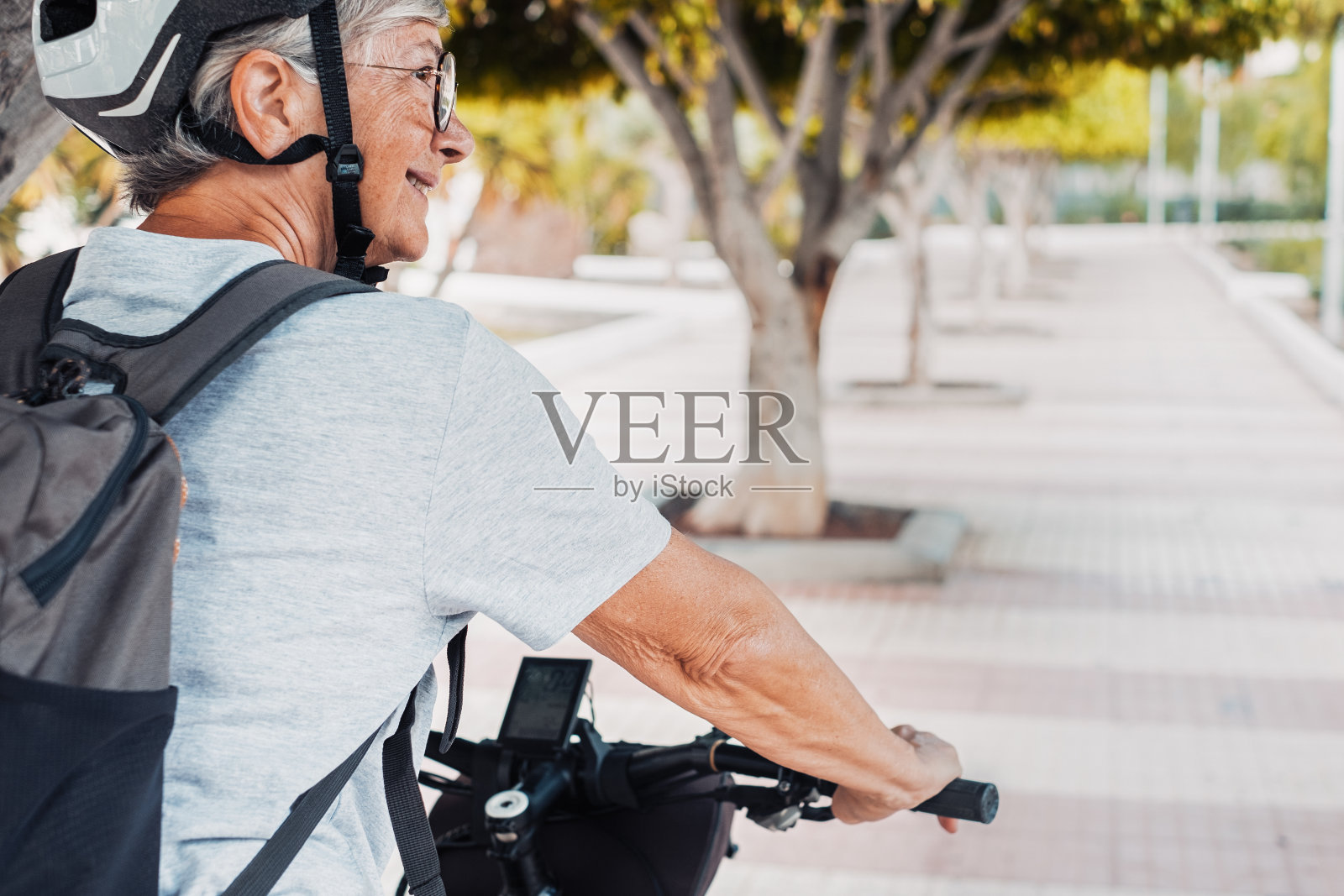 在城市公园的高级骑自行车的妇女戴着头盔和背包与她的电动自行车的背影。健康的生活方式和可持续的出行理念照片摄影图片