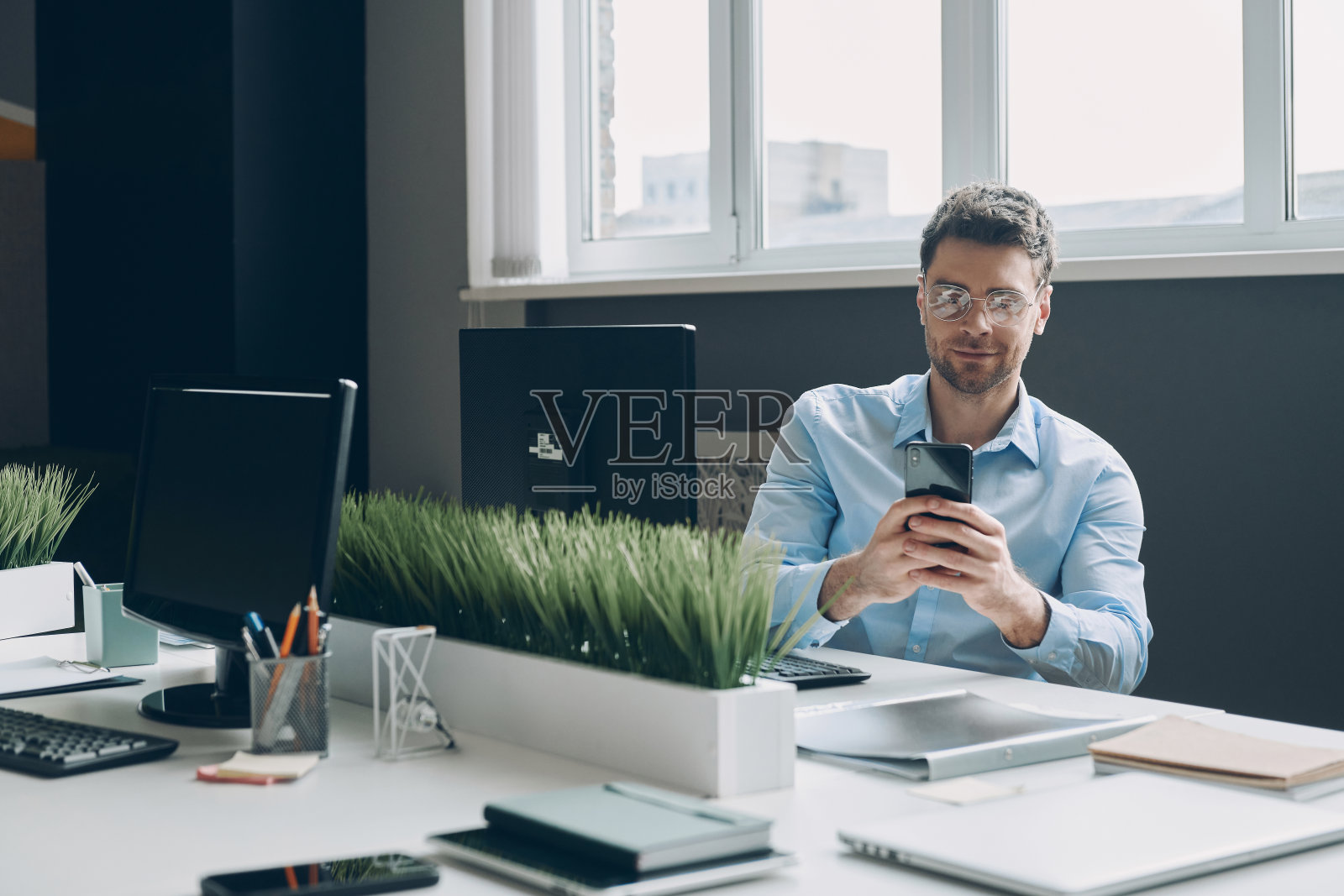 自信的年轻商人使用智能手机，而坐在他的工作地点在办公室照片摄影图片