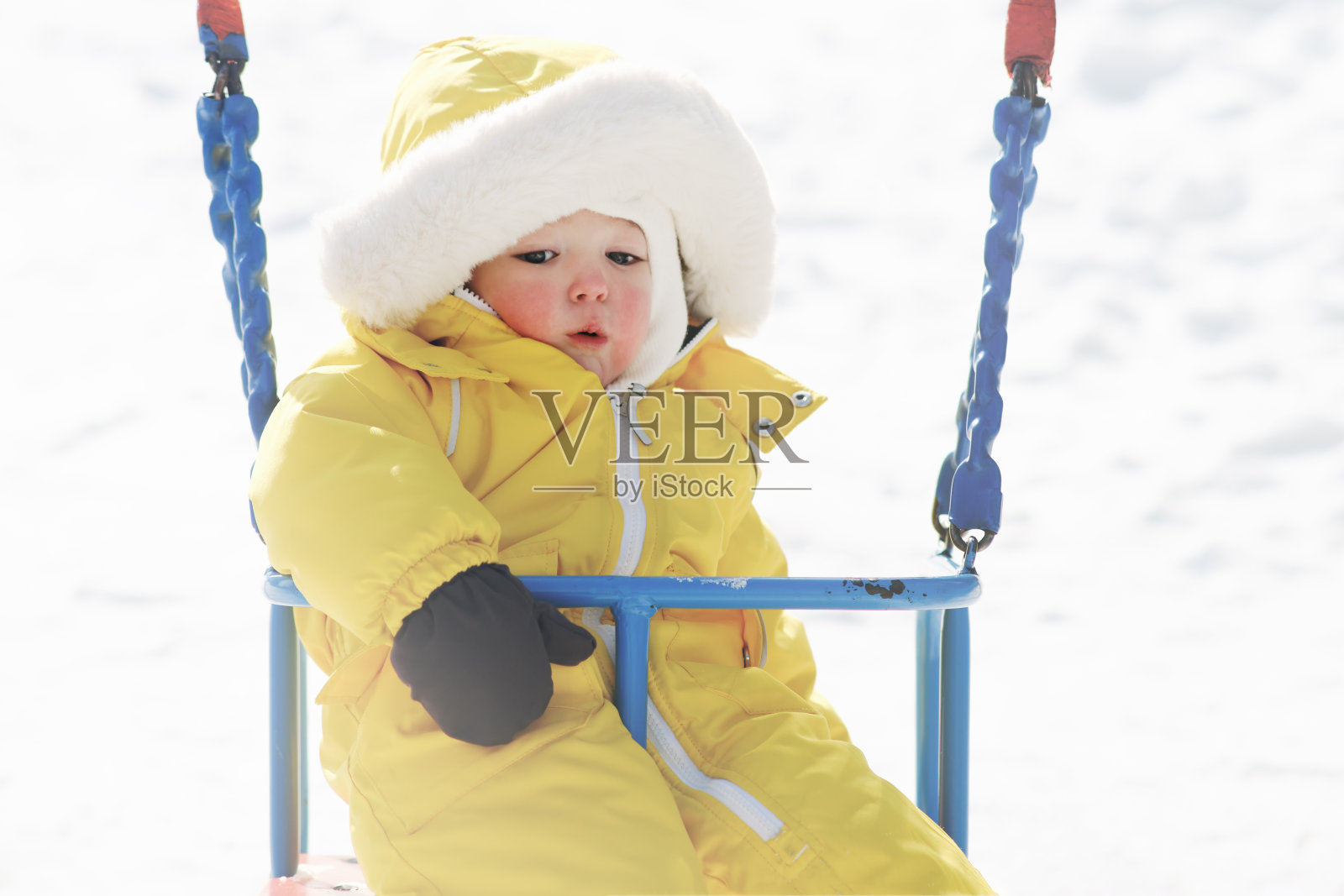 快乐的学步婴儿骑在秋千上的冬季操场。穿着黄色雪地服的小男孩在幼儿园荡秋千。孩子一岁八个月大照片摄影图片