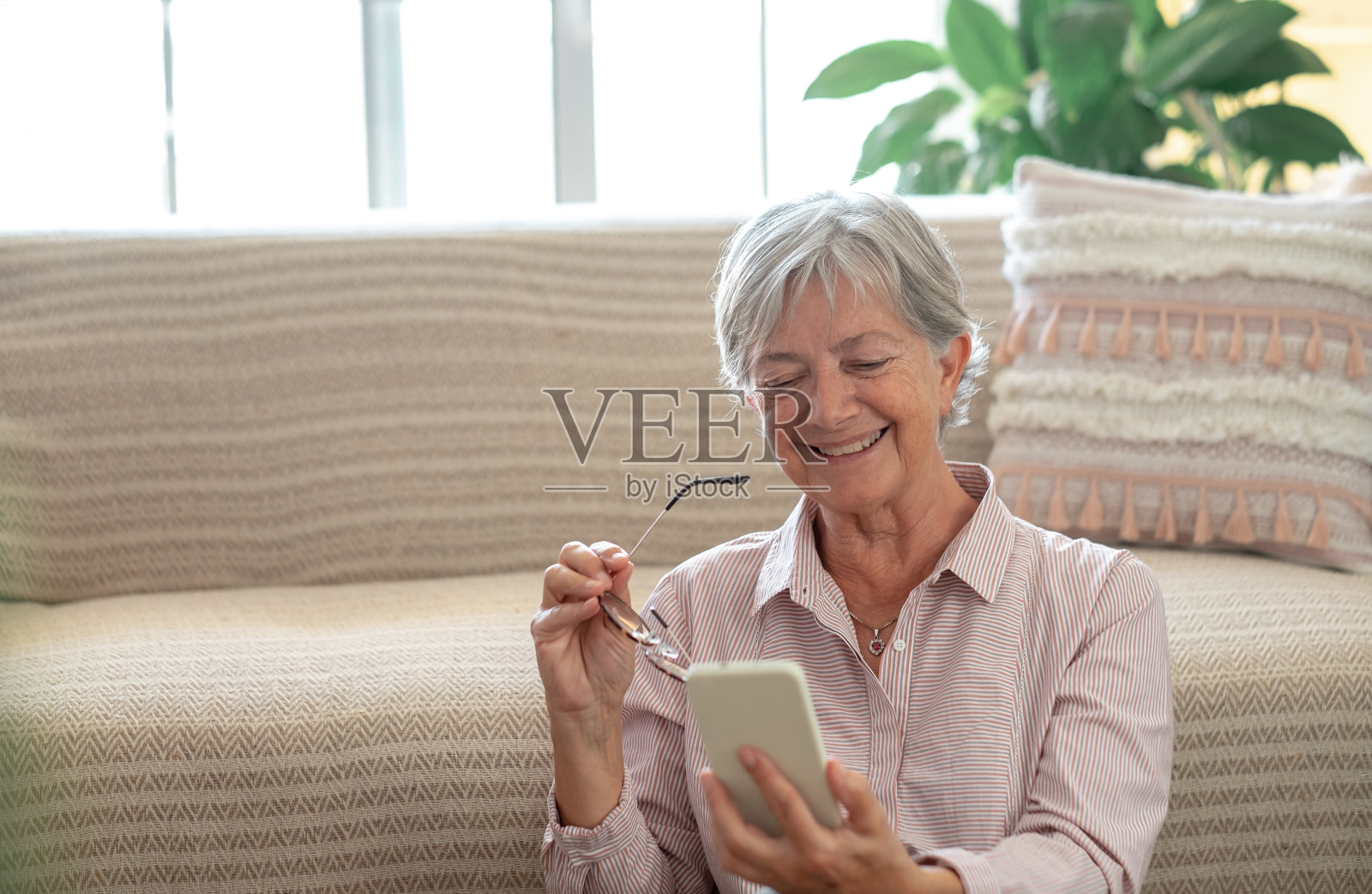 开朗的老年妇女放松地坐在家里的地板上，手里拿着智能手机看有趣的视频。微笑成熟的女士在家里使用移动应用程序。照片摄影图片