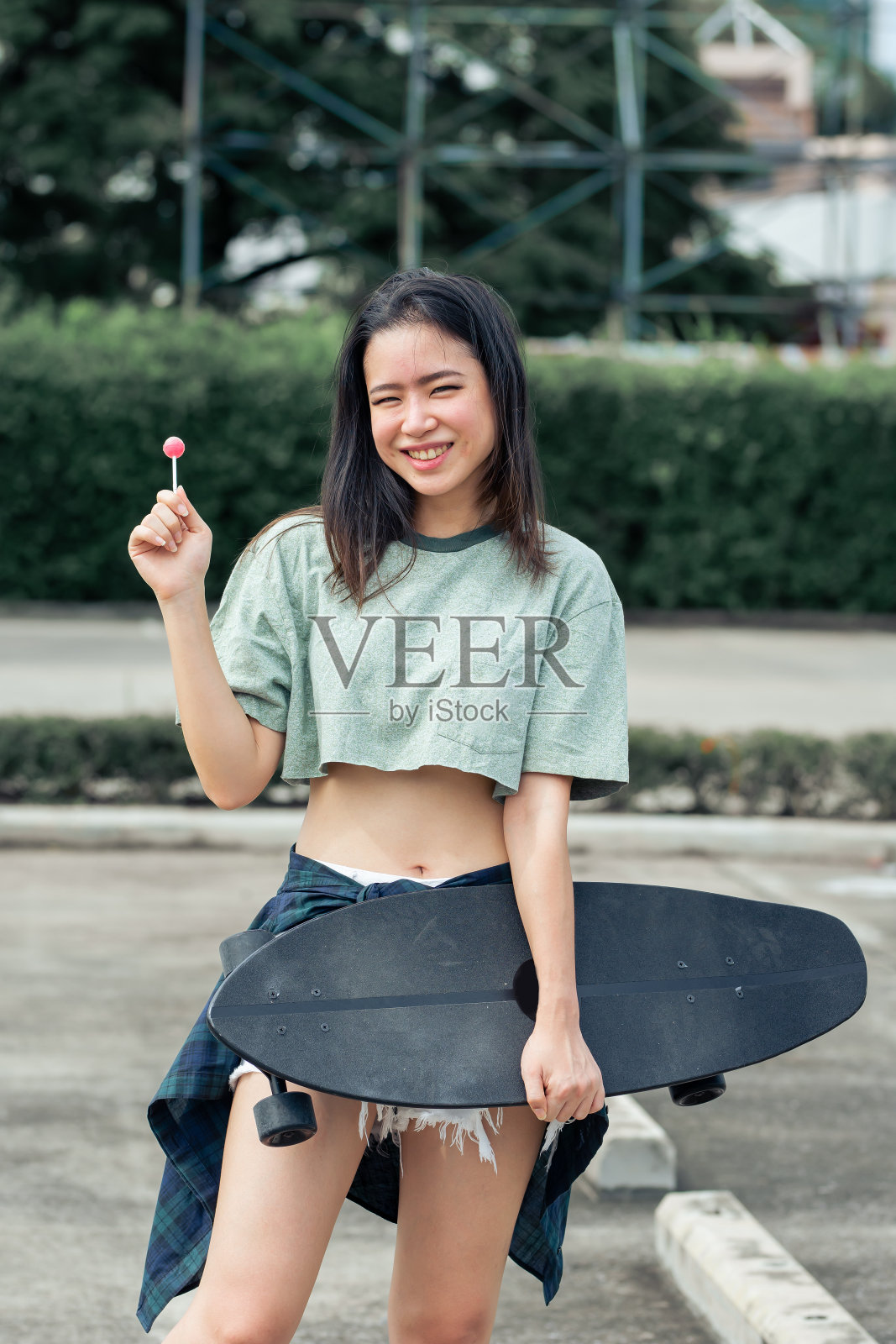 肖像美丽的体育亚洲女滑冰穿着时髦的衣服与棒棒糖，微笑与幸福，拿着滑板，站在户外与复制空间。活动和冒险概念。照片摄影图片