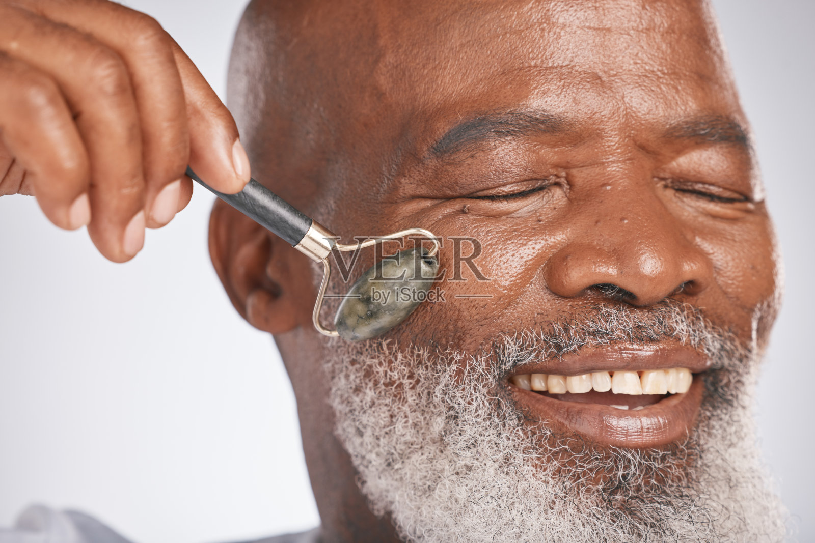 面部滚轮，老年男子和皮肤按摩和为护肤，自我护理和皮肤美容的工具。一个快乐的黑人男性的头部与玫瑰石英灰色工作室背景抗衰老照片摄影图片