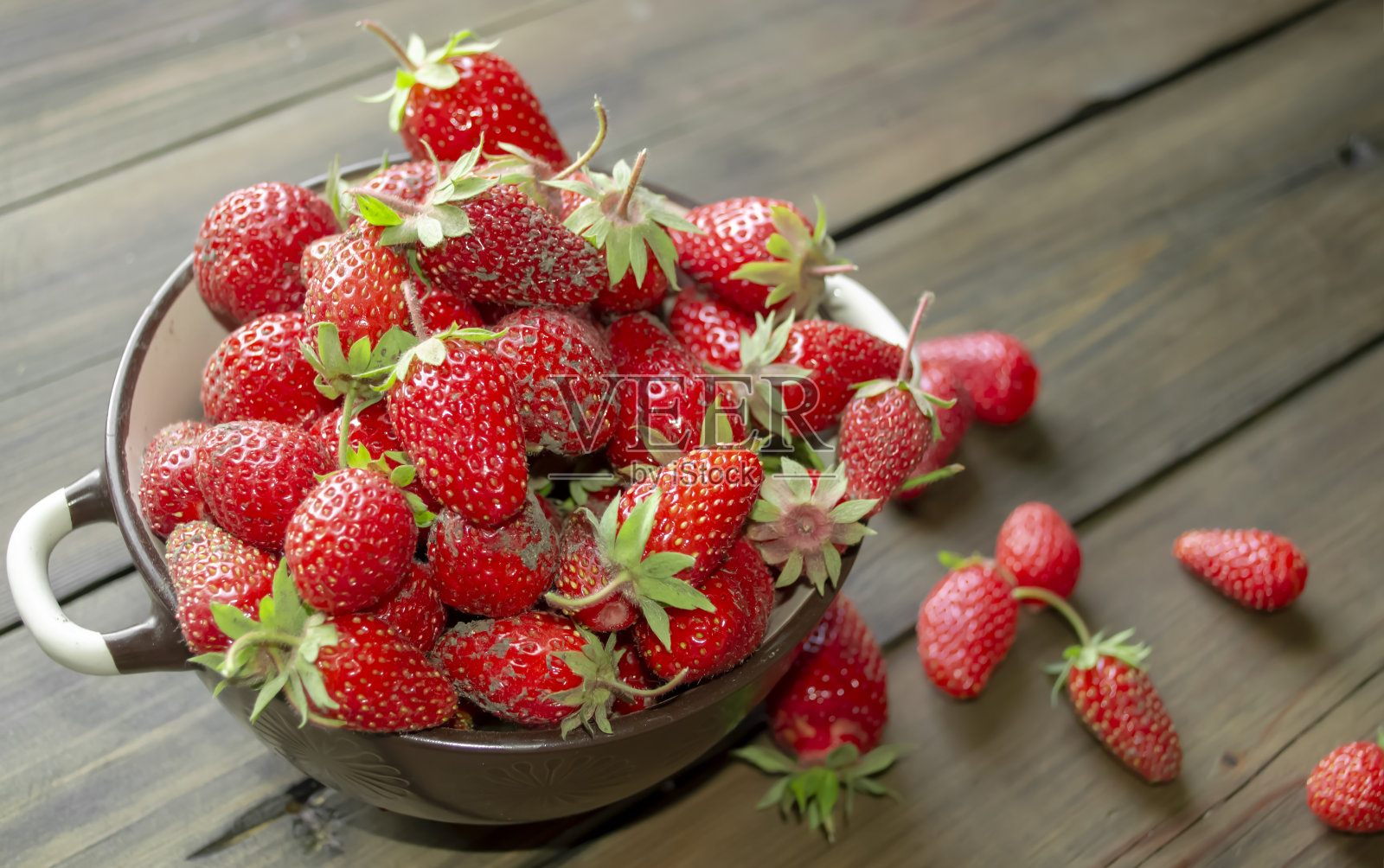 装在盘子里的草莓。新鲜的春天浆果——从花园里摘的草莓放在盘子里和桌子上。在一个深盘子里有脏草莓和绿色的尾巴。照片摄影图片