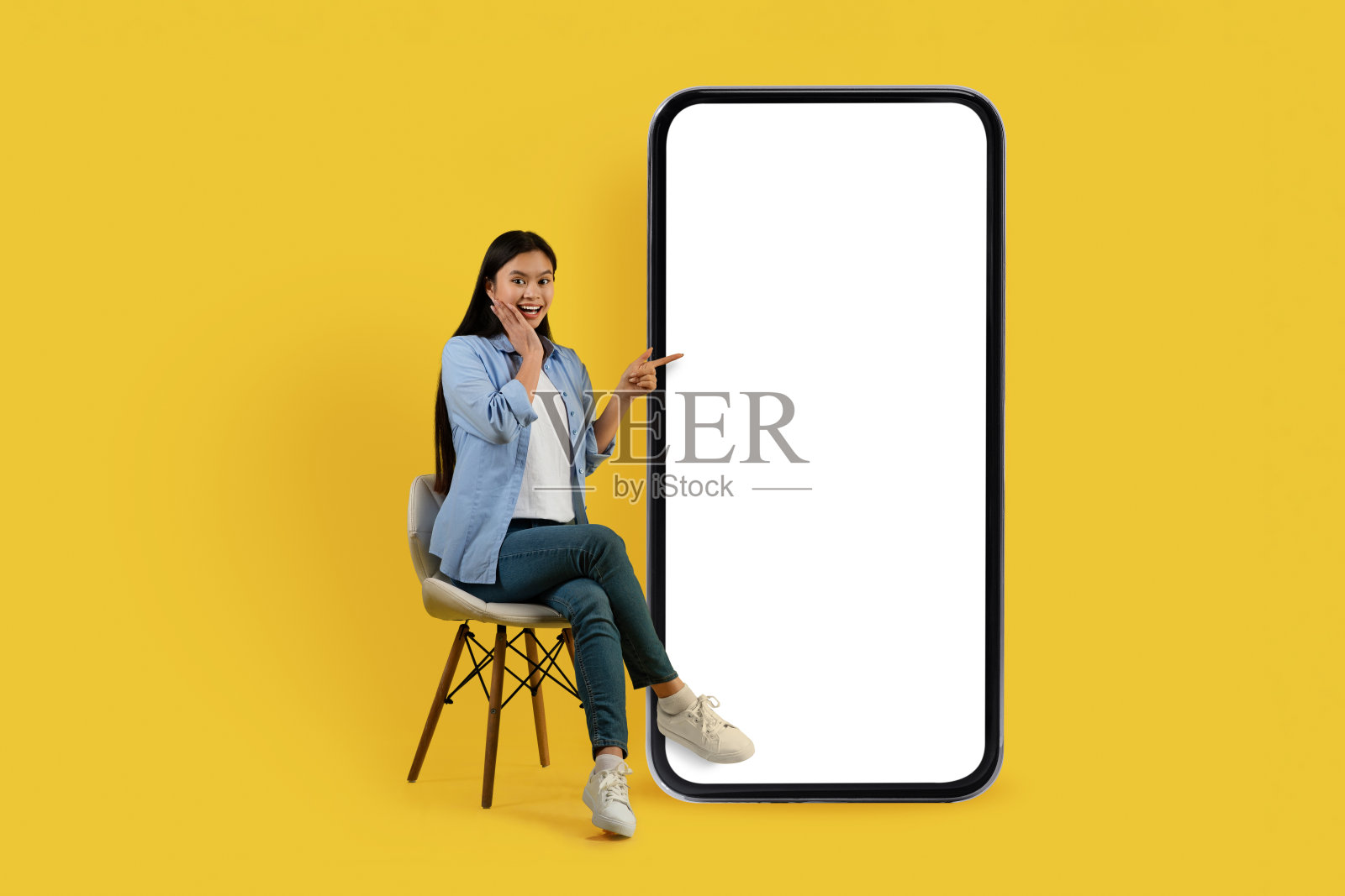 微笑震惊的年轻中国女学生在休闲坐在椅子上，手指着巨大的智能手机与空屏幕照片摄影图片