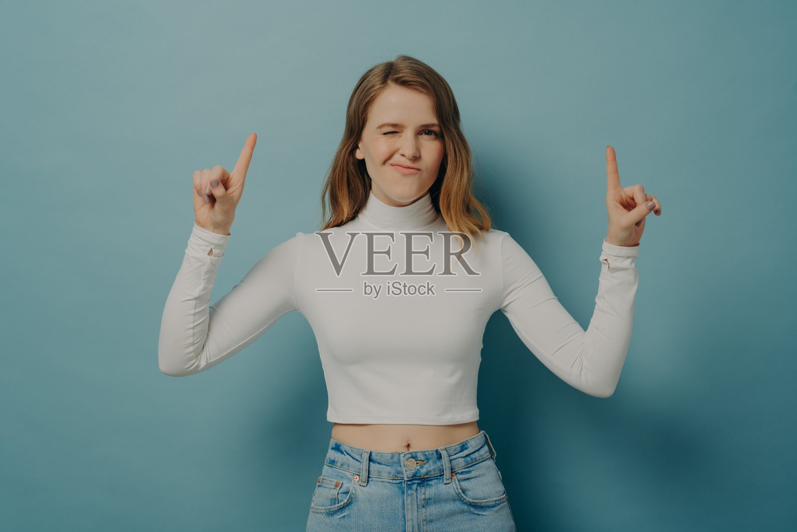 千禧一代的女孩用食指向上指，对着相机眨眼照片摄影图片